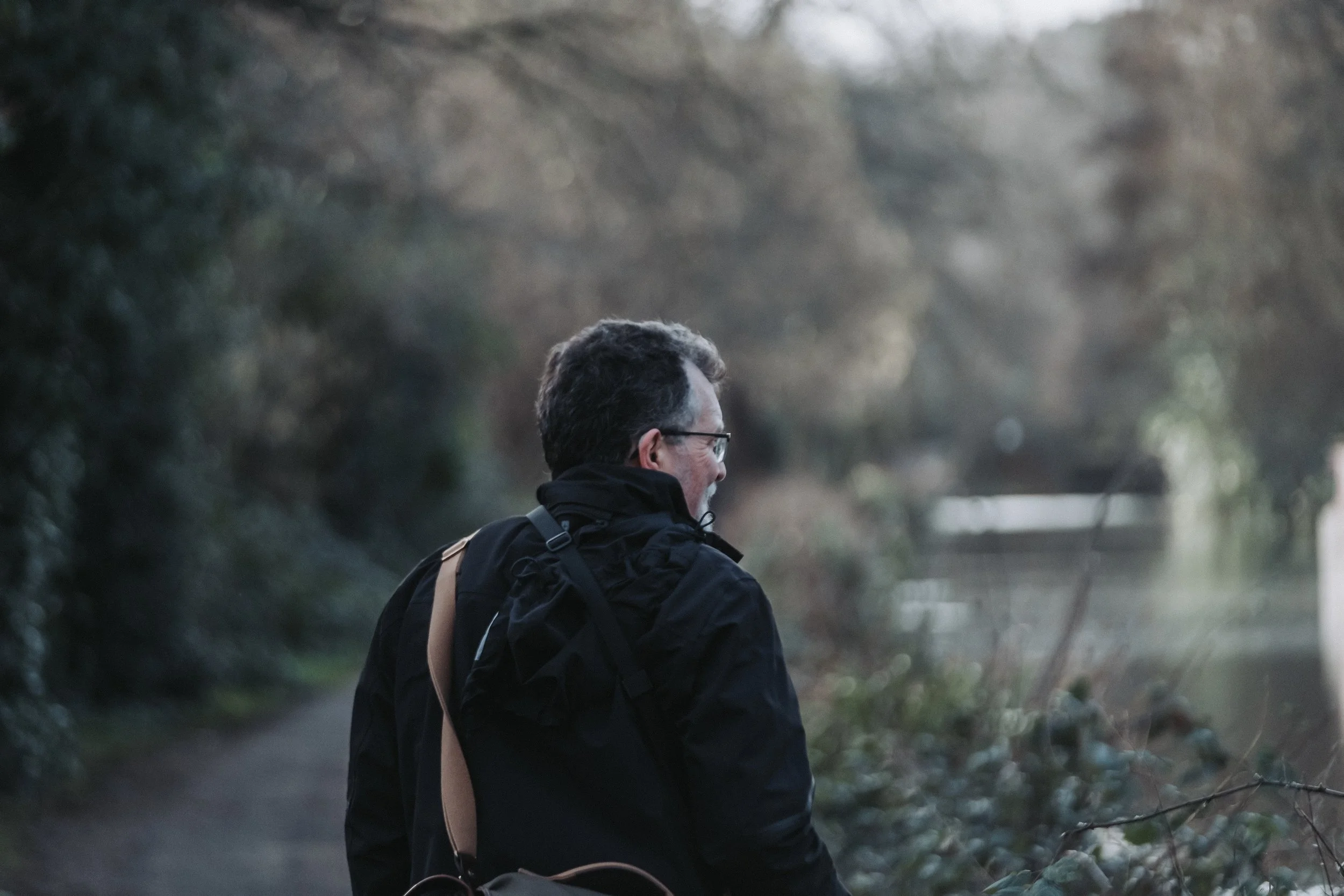 A man with glasses and brown hair wearing a black jacket and a beige strap bag, walking along a trail by a river in a wooded area during dusk or dawn.