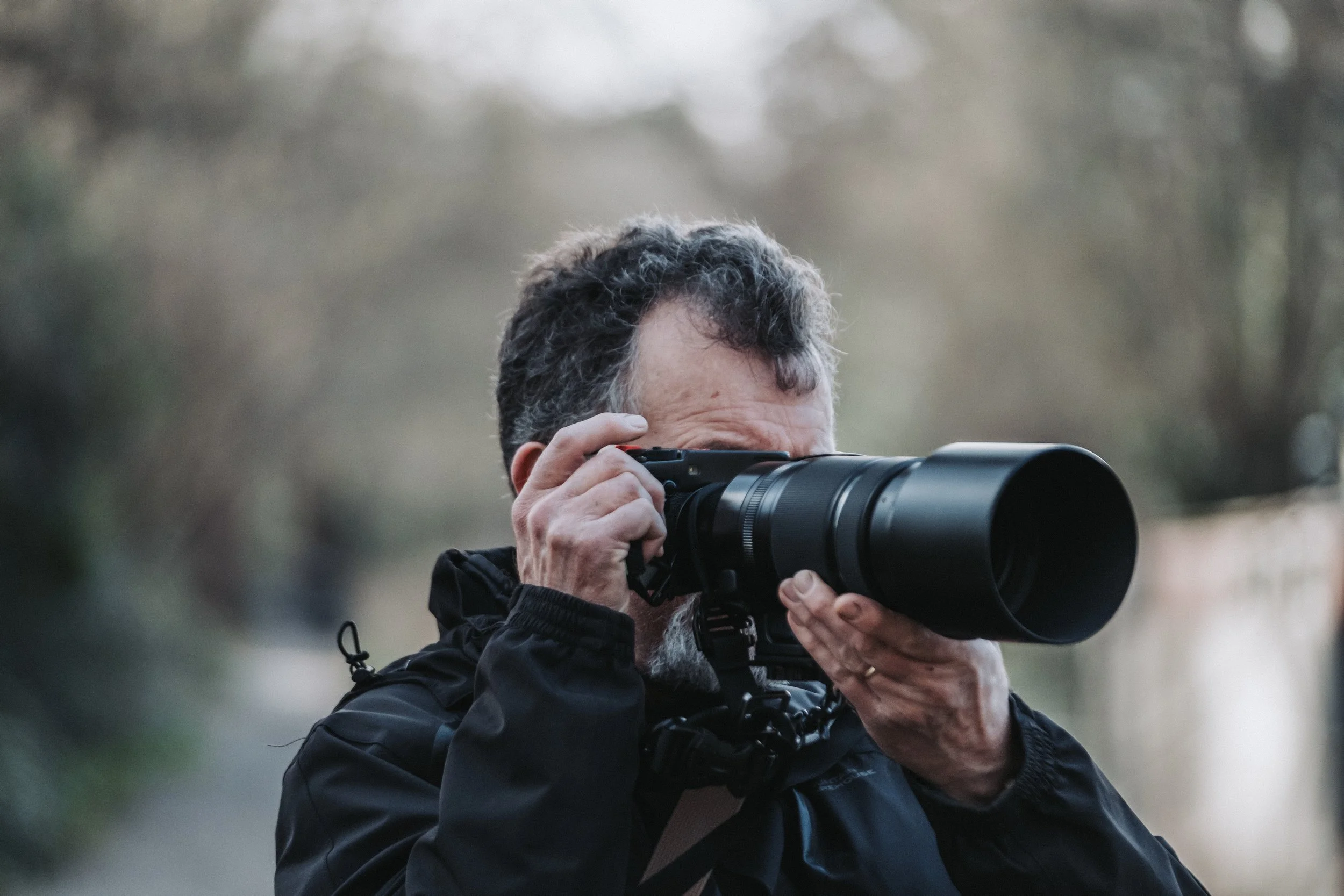 A man with dark curly hair is looking through a large camera with a telephoto lens, wearing a black jacket outdoors.