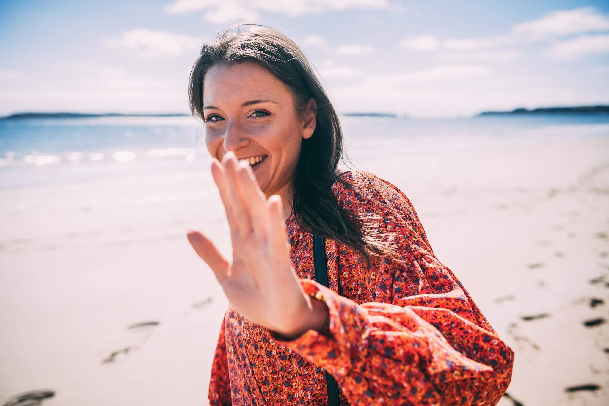 A woman smiling and waving at the beach during daytime, wearing a red patterned jacket.