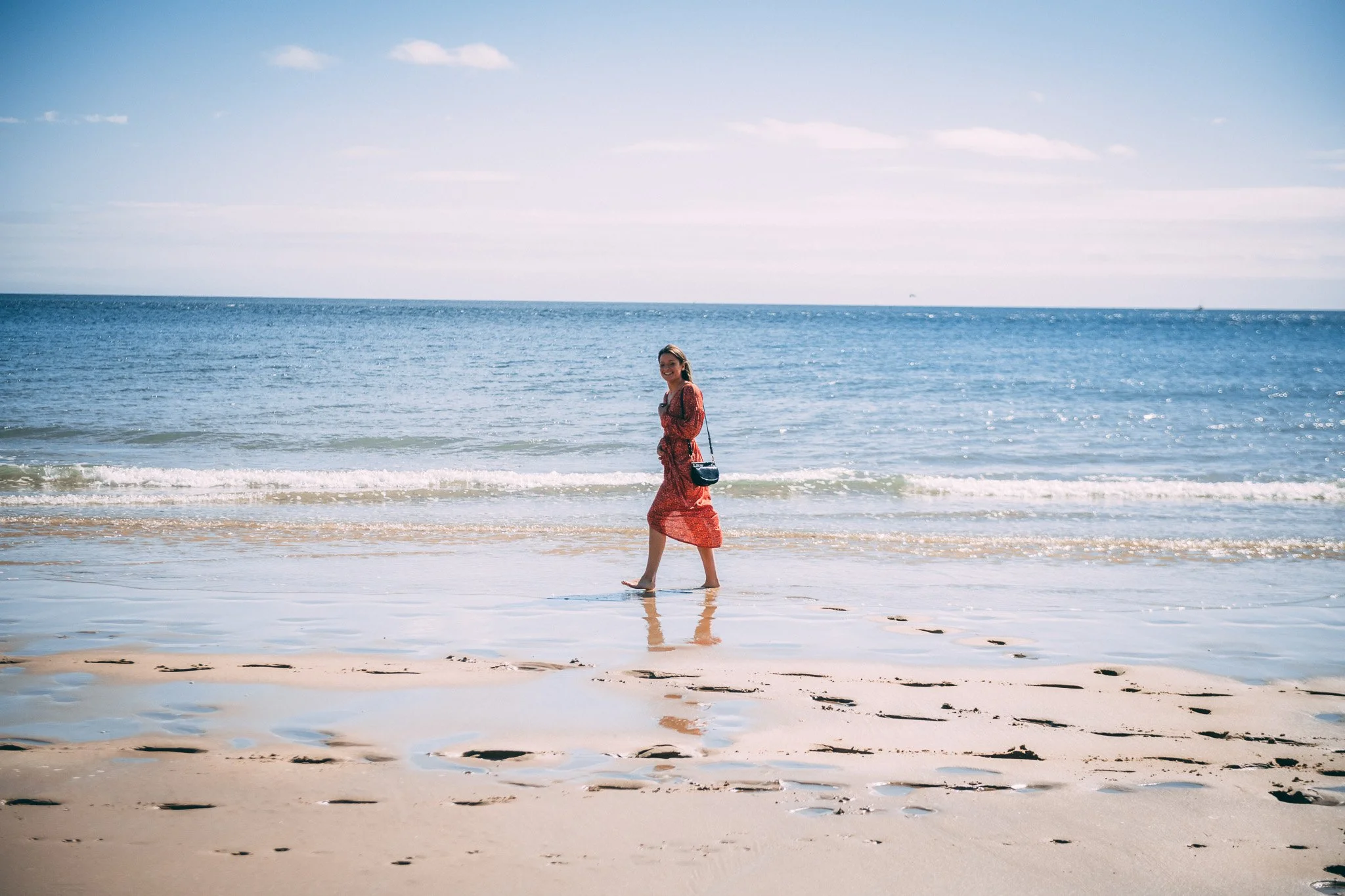 A woman in a red dress walking along the shoreline of a beach, with gentle waves and a clear blue sky in the background.