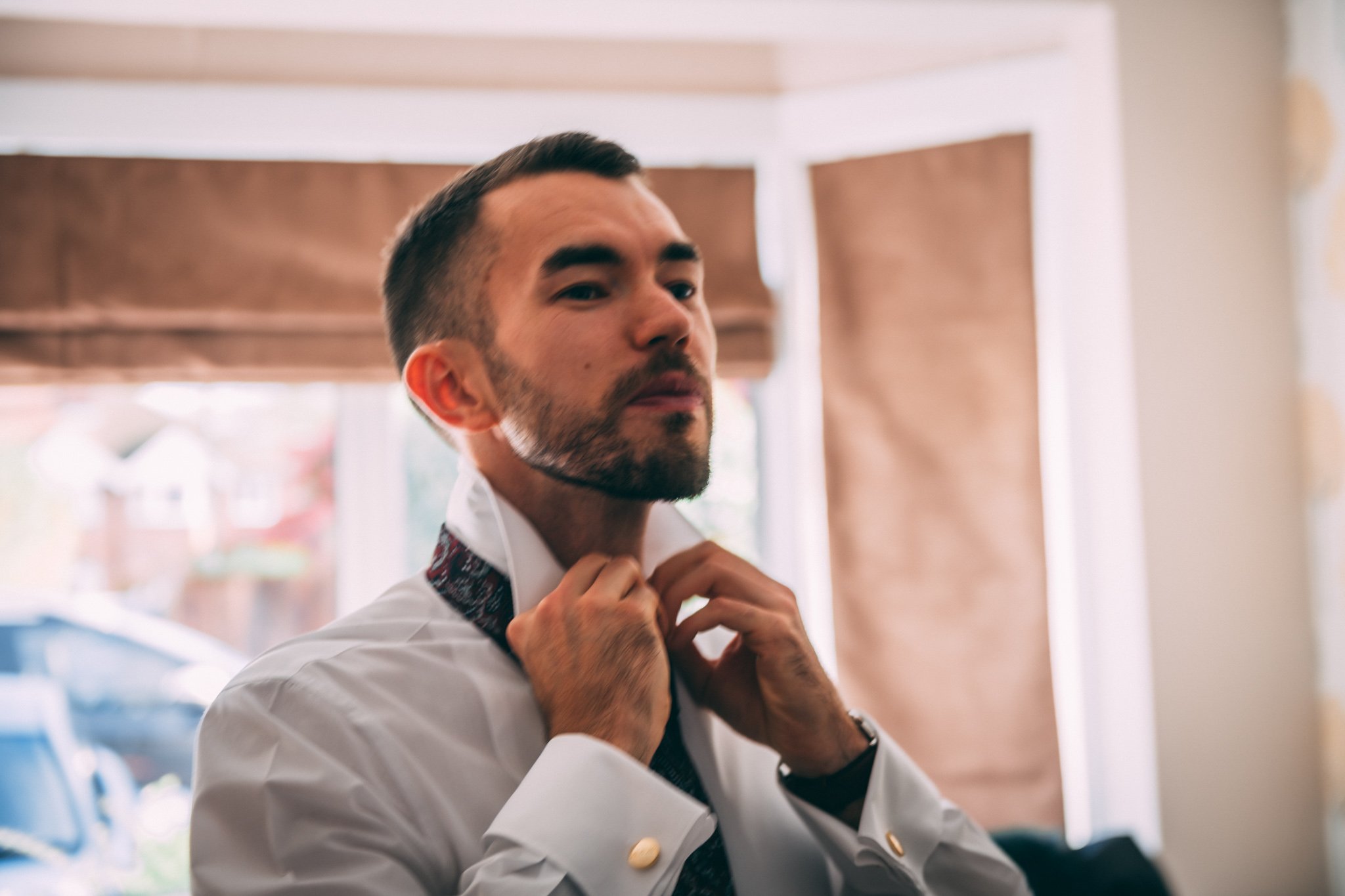 A man adjusting his necktie in a well-lit room with large windows and brown curtains.