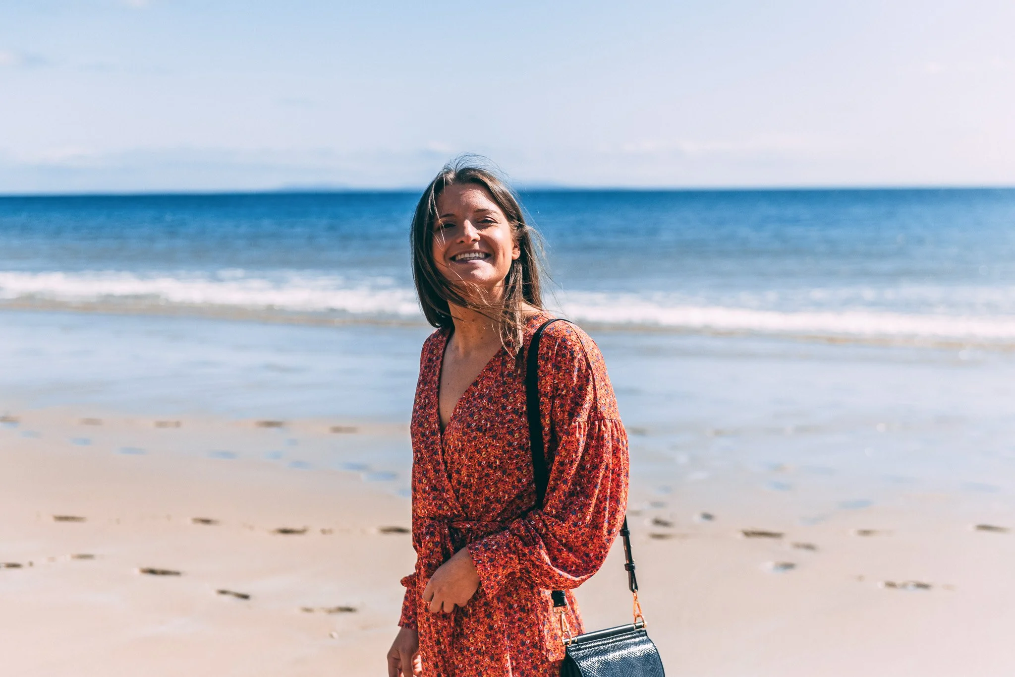 Woman smiling in a red floral dress on the beach with ocean waves in the background.