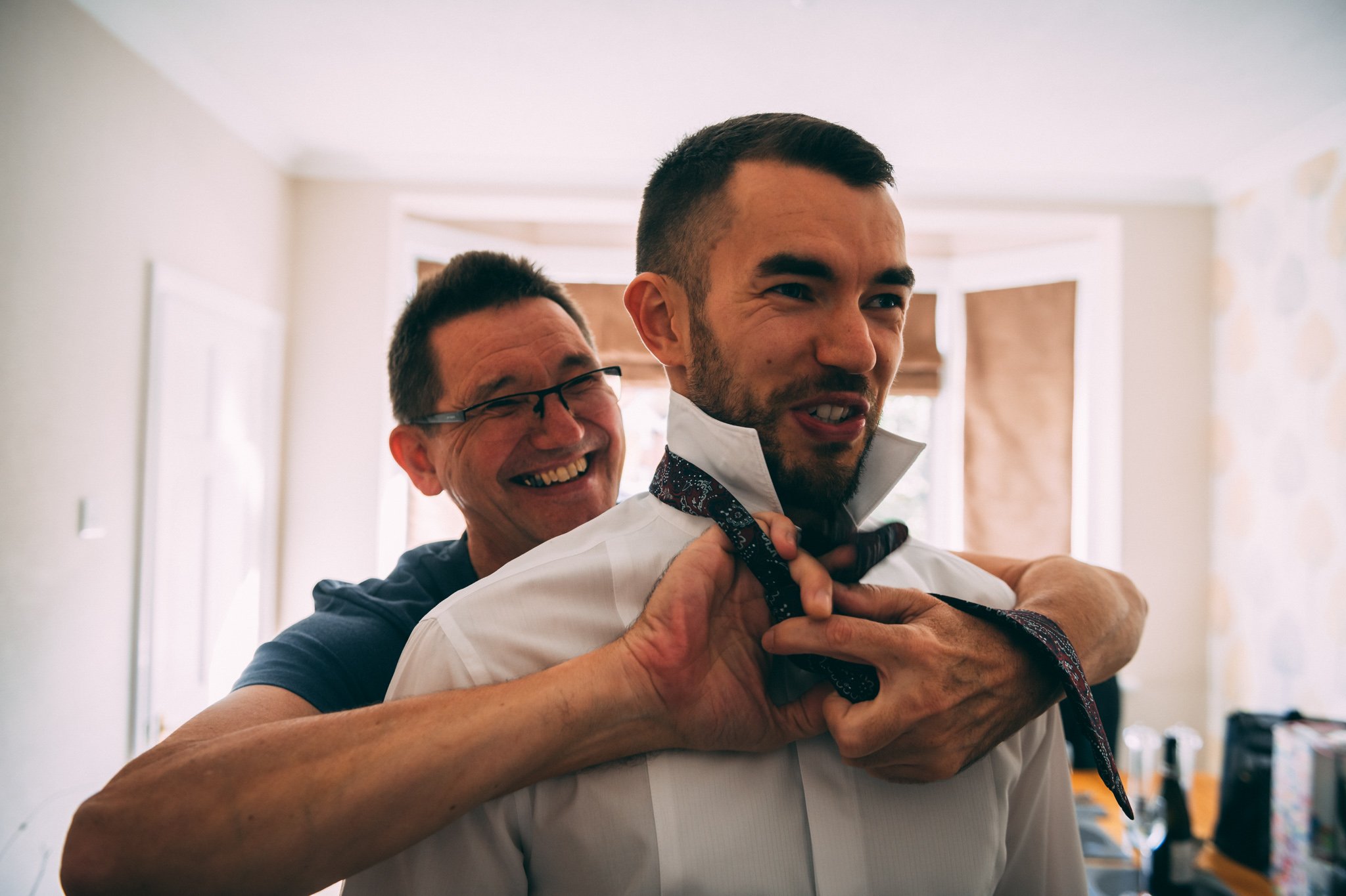 Two men preparing for a formal event, one helping the other tighten his tie in a bright room with large windows.