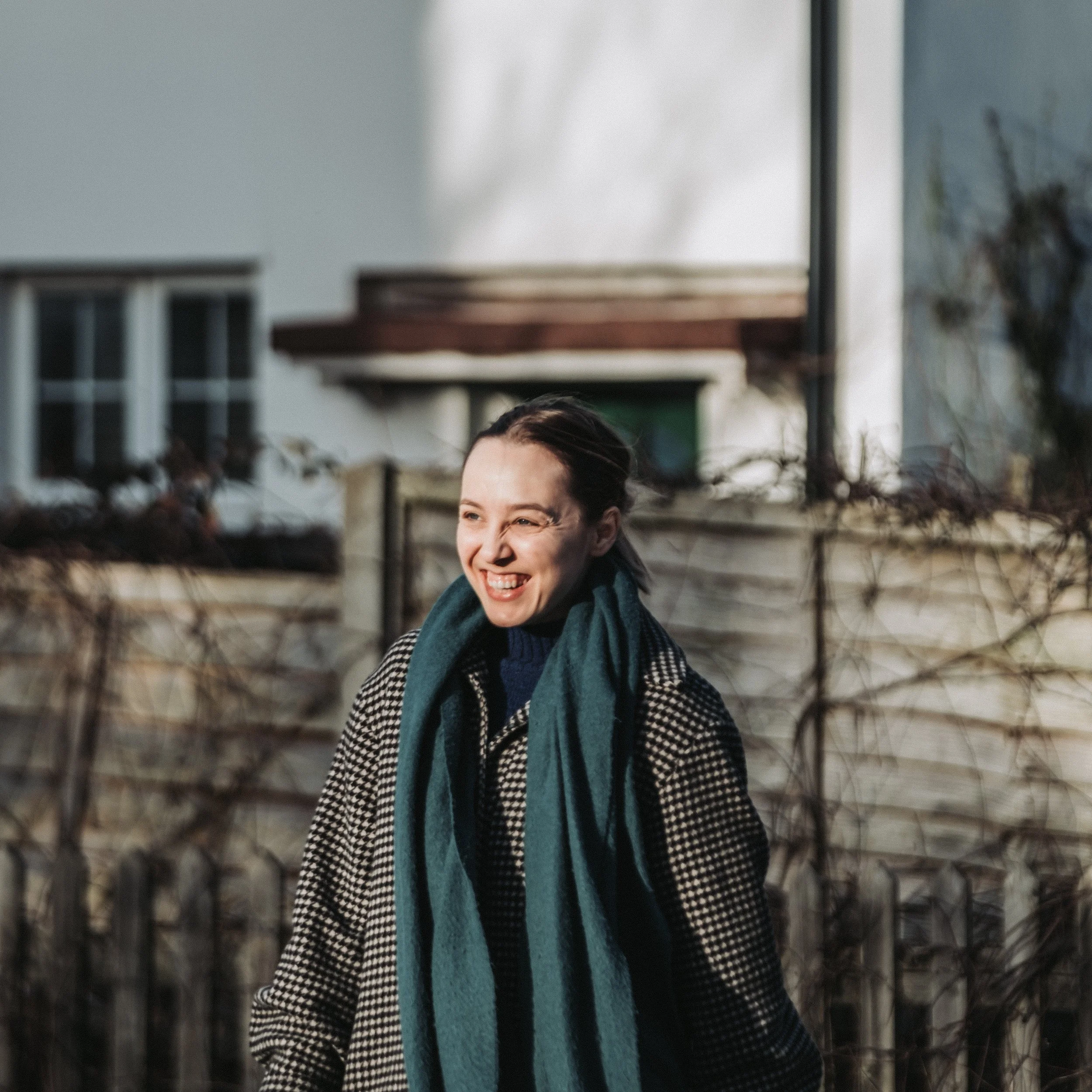 A woman with dark hair pulled back, smiling and squinting in sunlight, wearing a black and white checkered coat and teal scarf outdoors, with a wooden fence and house in the background.
