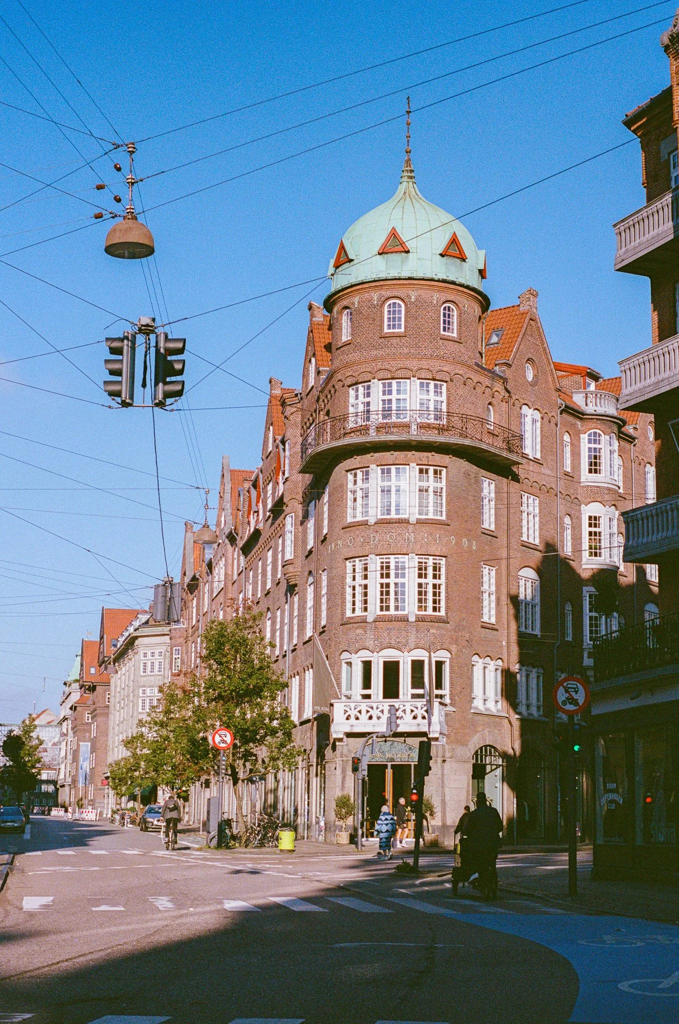 A street scene with a historic brick building featuring a rounded tower with a green dome roof. The building has multiple windows and balconies. There are street lights, overhead tram wires, pedestrians, and bicycles on the sidewalk, with a clear blu