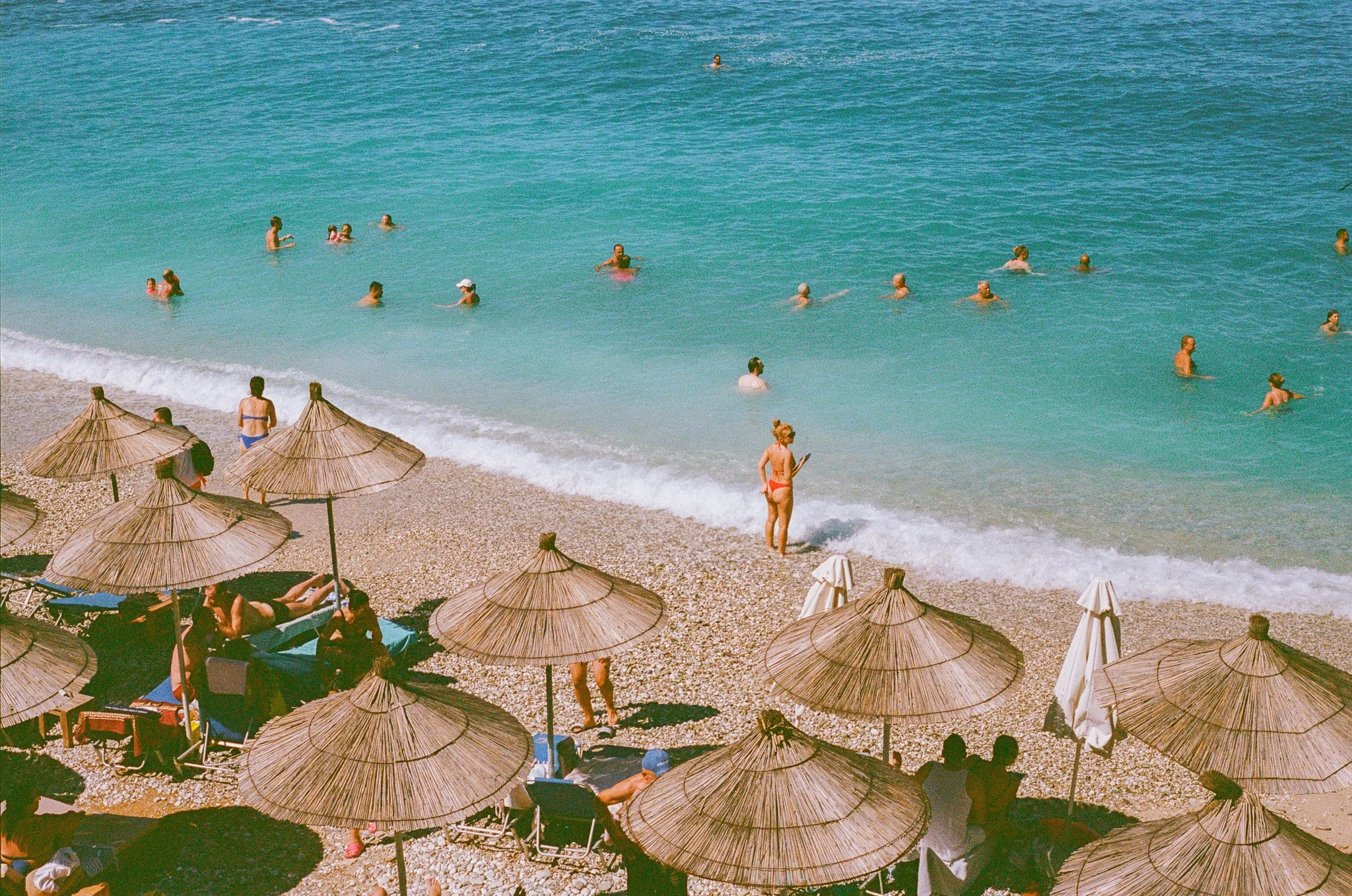 People swimming and relaxing at a beach with straw umbrellas and pebbled shore.