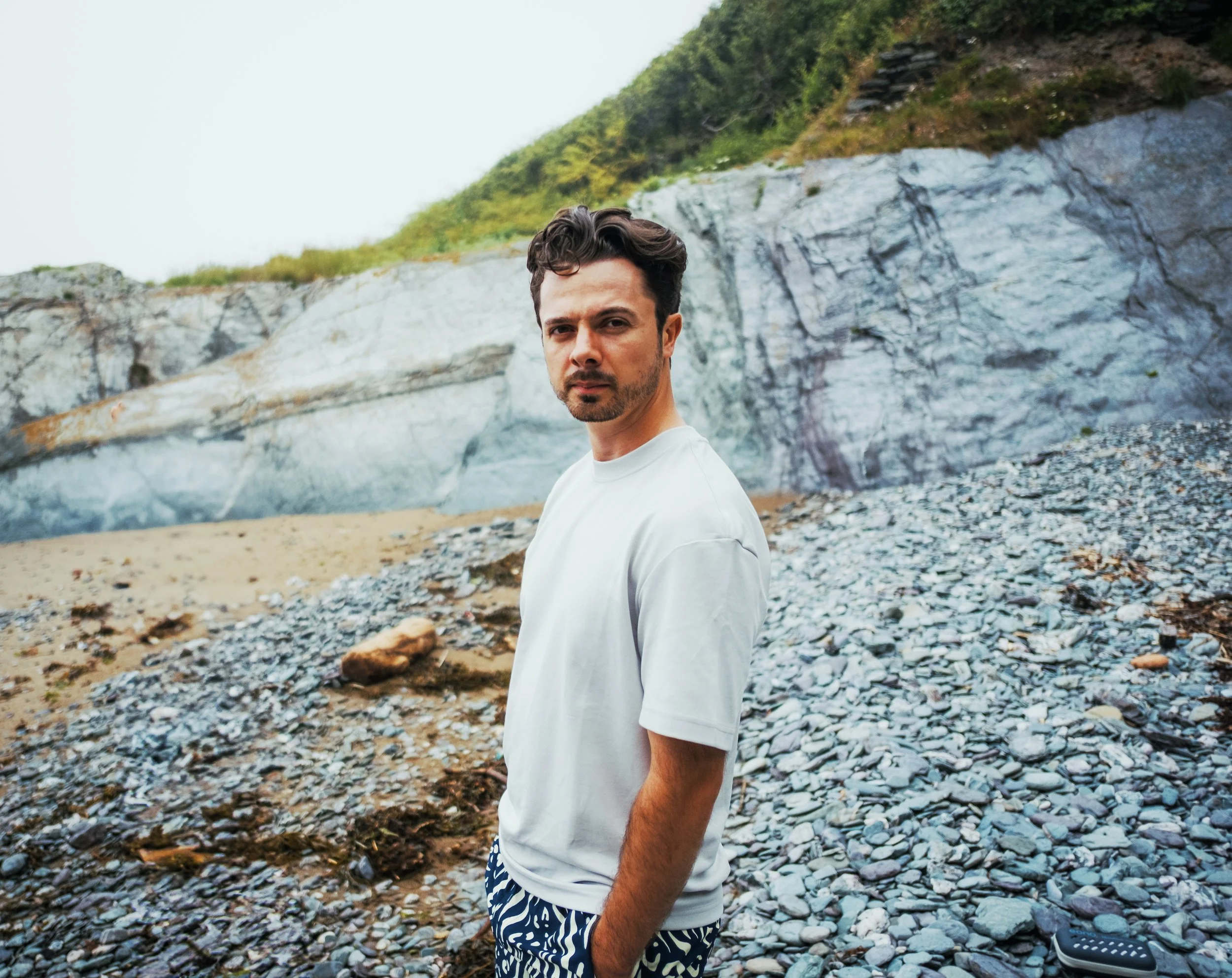A man with dark hair and a beard stands on a rocky beach with steep cliffs behind him and greenery on top. He is wearing a white t-shirt and patterned shorts, looking at the camera with a serious expression.