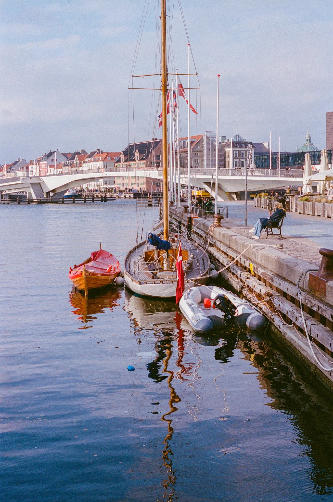 Boats docked along a waterfront promenade with a man sitting on a bench, a modern bridge in the background, and a city skyline with buildings and a dome on the horizon.