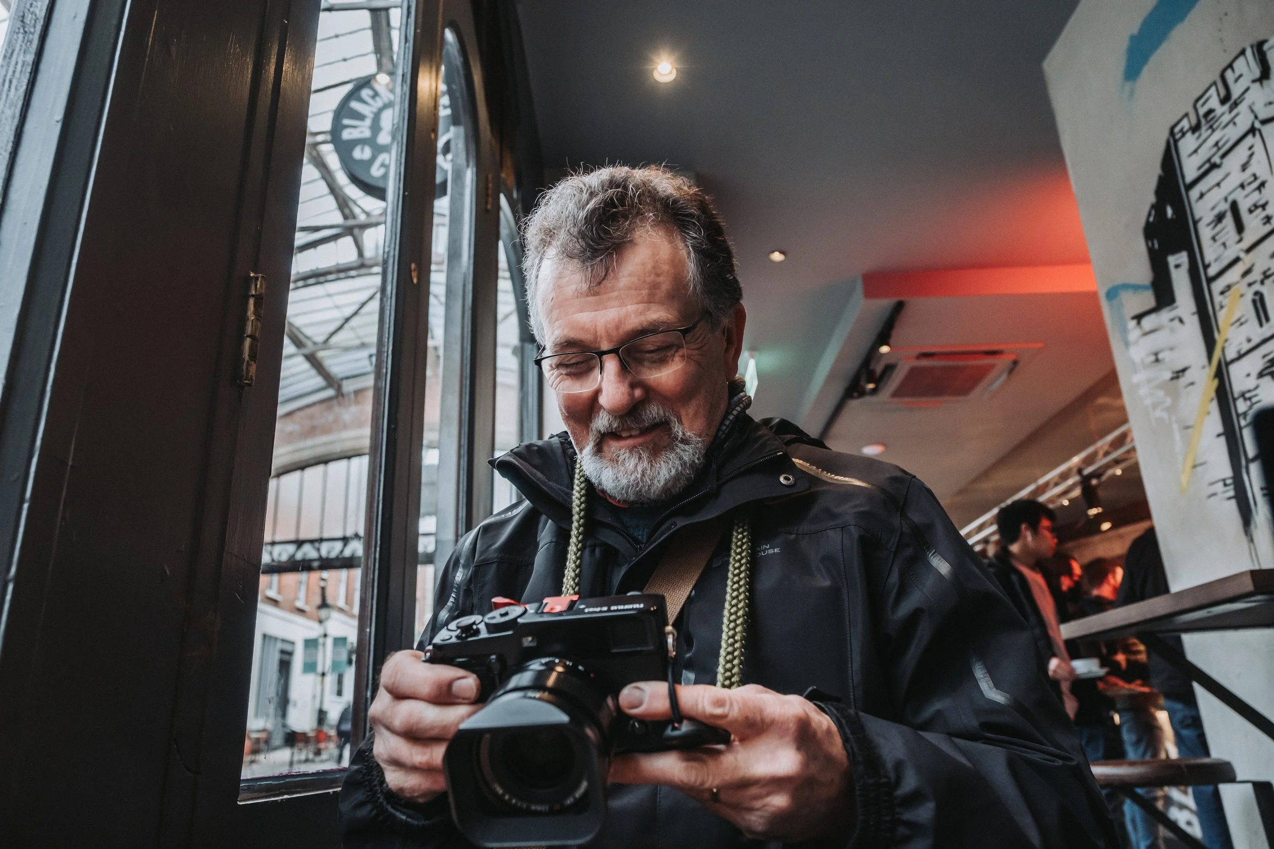 A man with glasses and a gray beard looks at a camera inside a cafe or restaurant.