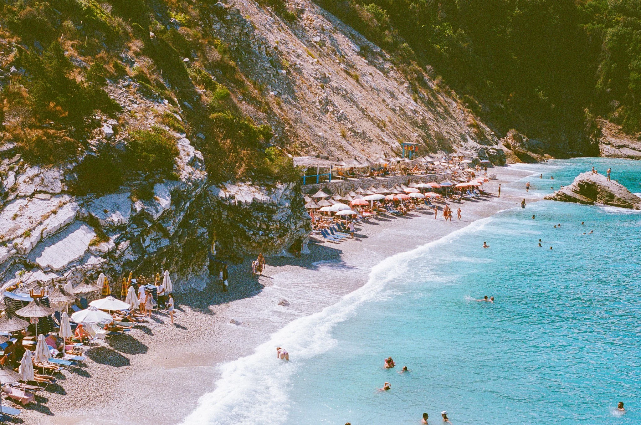 A beach scene with turquoise water, people swimming and wading, rocky cliffs, and a shoreline with umbrellas and lounge chairs.