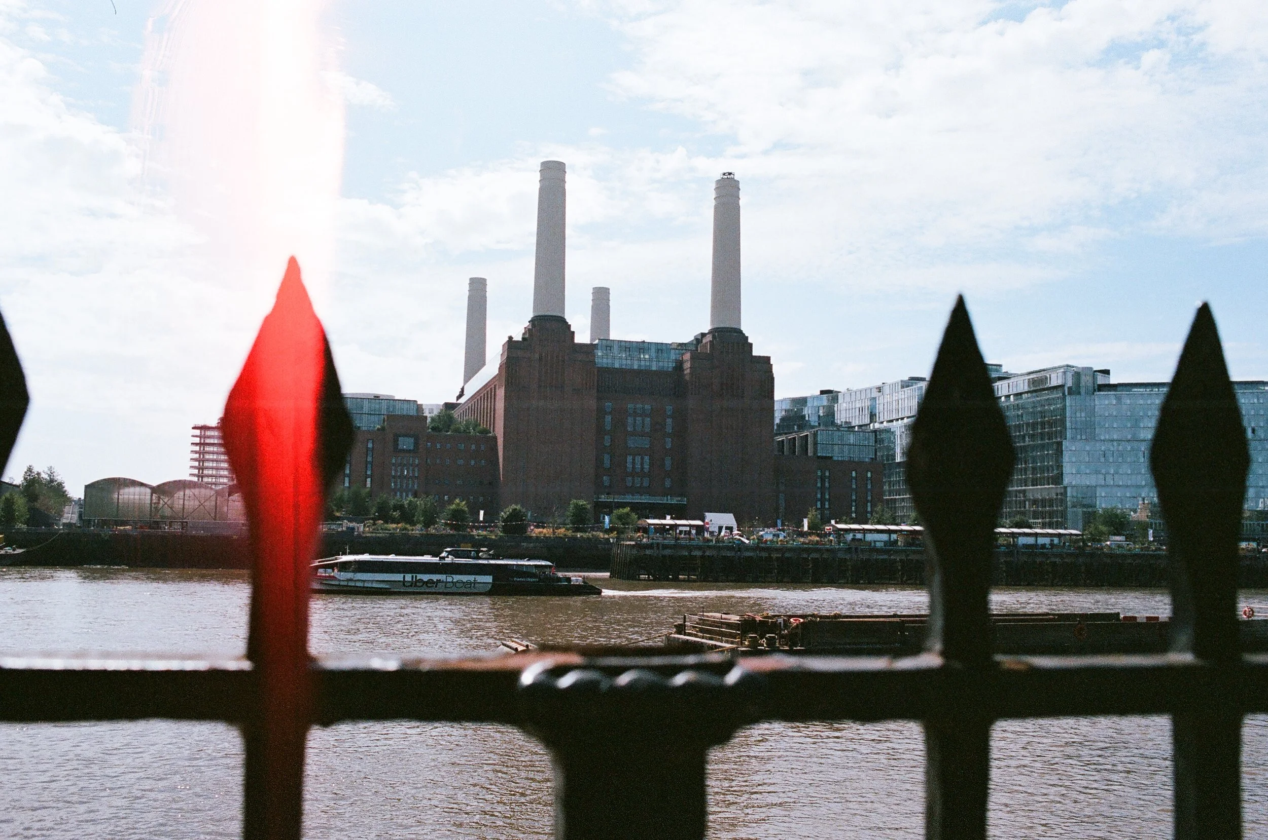 View of a large industrial building with tall white chimneys across a river, seen through a black metal fence with pointed tips and one red tip, and a boat named 'Uber Boat' on the water.