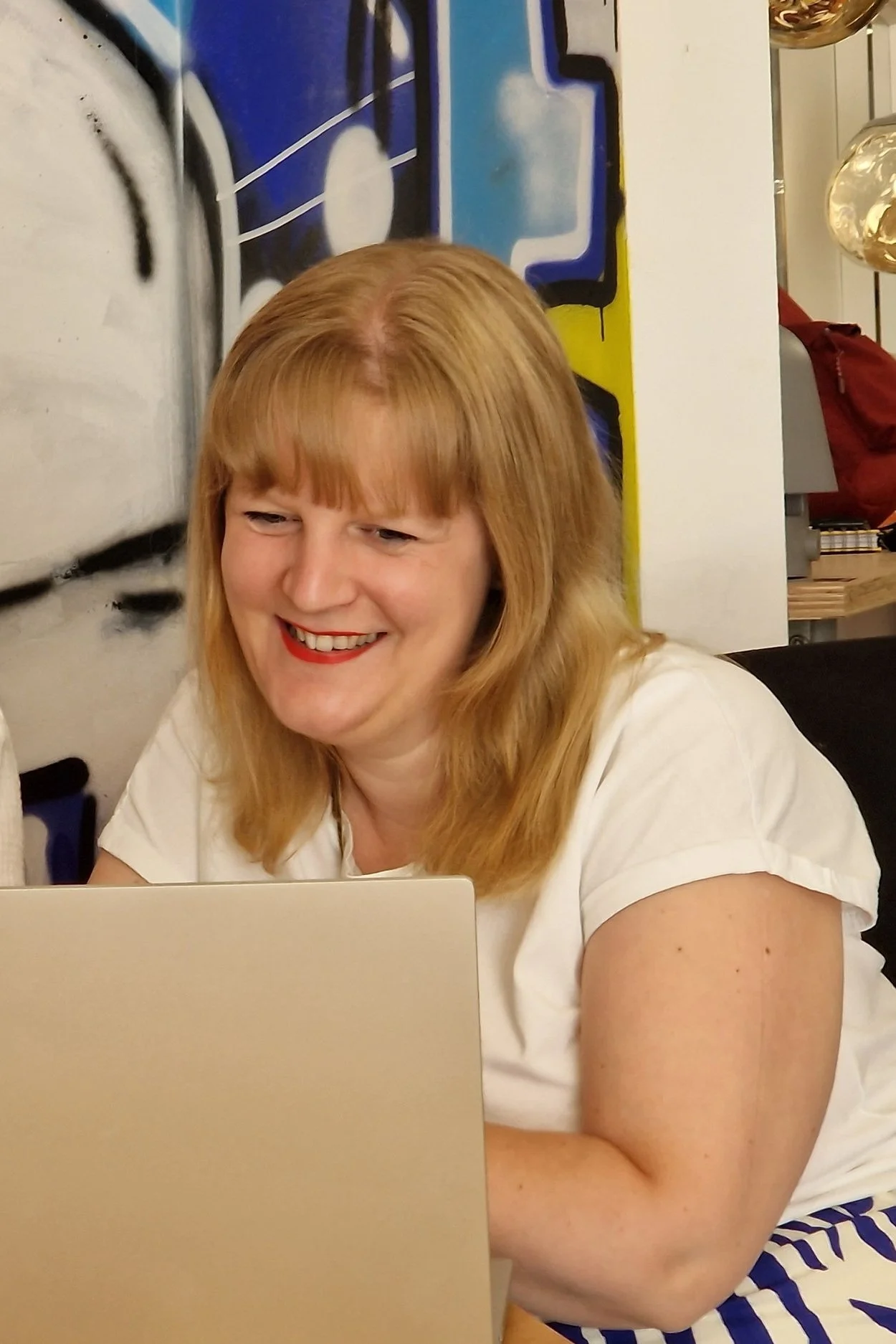 A woman with blonde hair smiling while looking at a laptop, seated in front of a colorful graffiti-style wall.