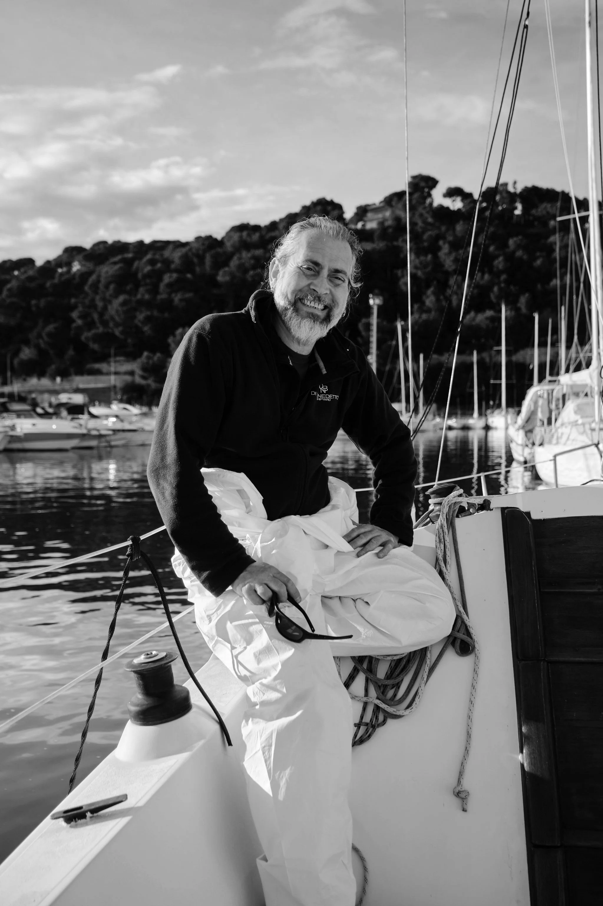 A man with long hair and a beard, crossed arms, wearing a T-shirt and dark pants, standing against a plain wall.