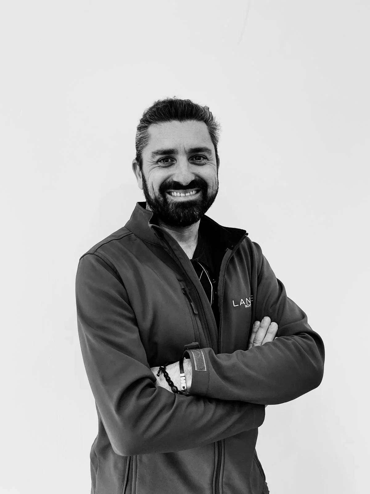 A smiling man with a beard and styled hair, wearing a zip-up jacket and crossing his arms, standing against a plain white wall.