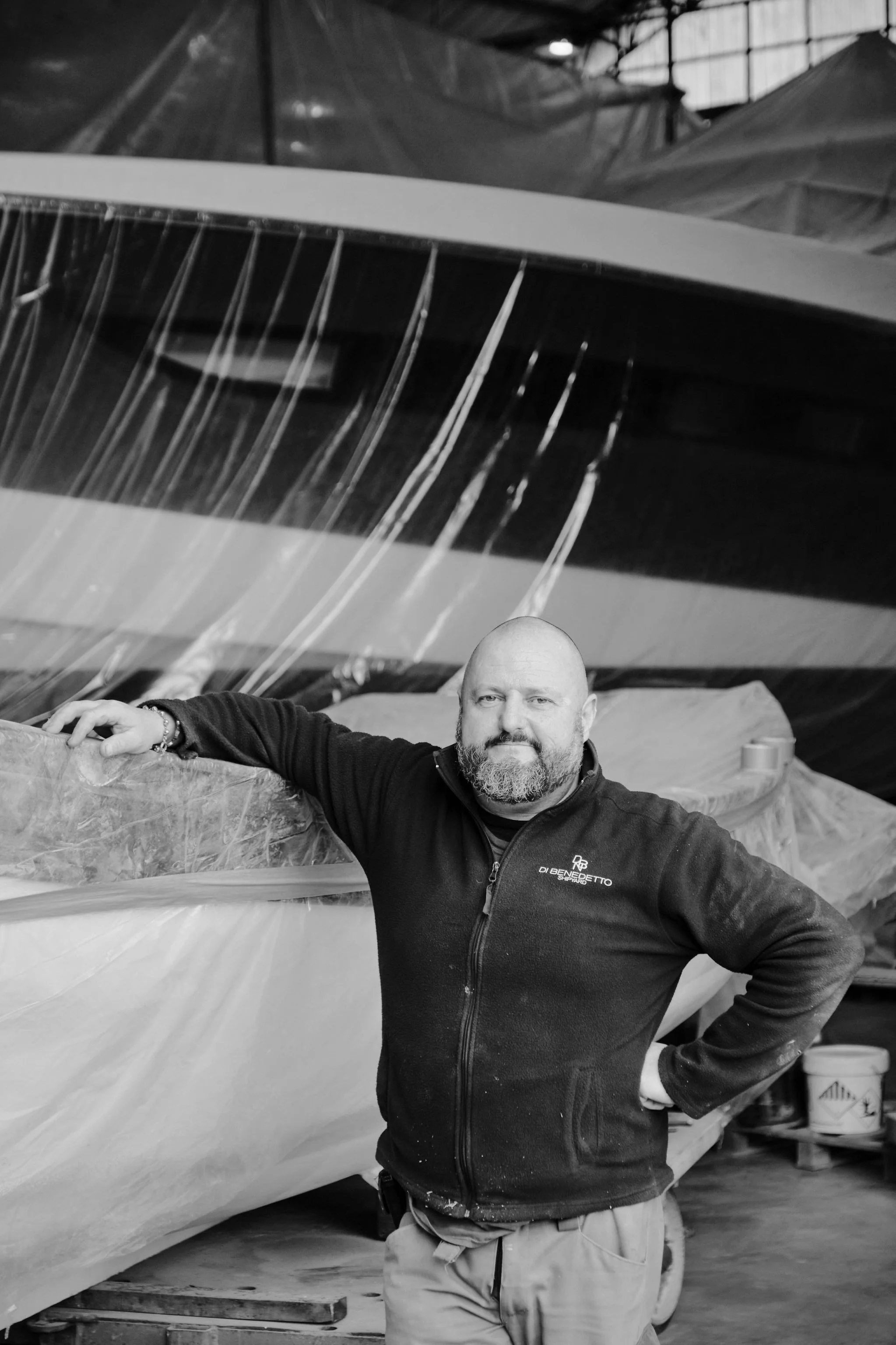 Black and white photo of a bald man with a beard, crossing his arms, wearing a zip-up jacket with a logo that reads 'LAND'S YACHTING' on the chest, standing against a plain background.