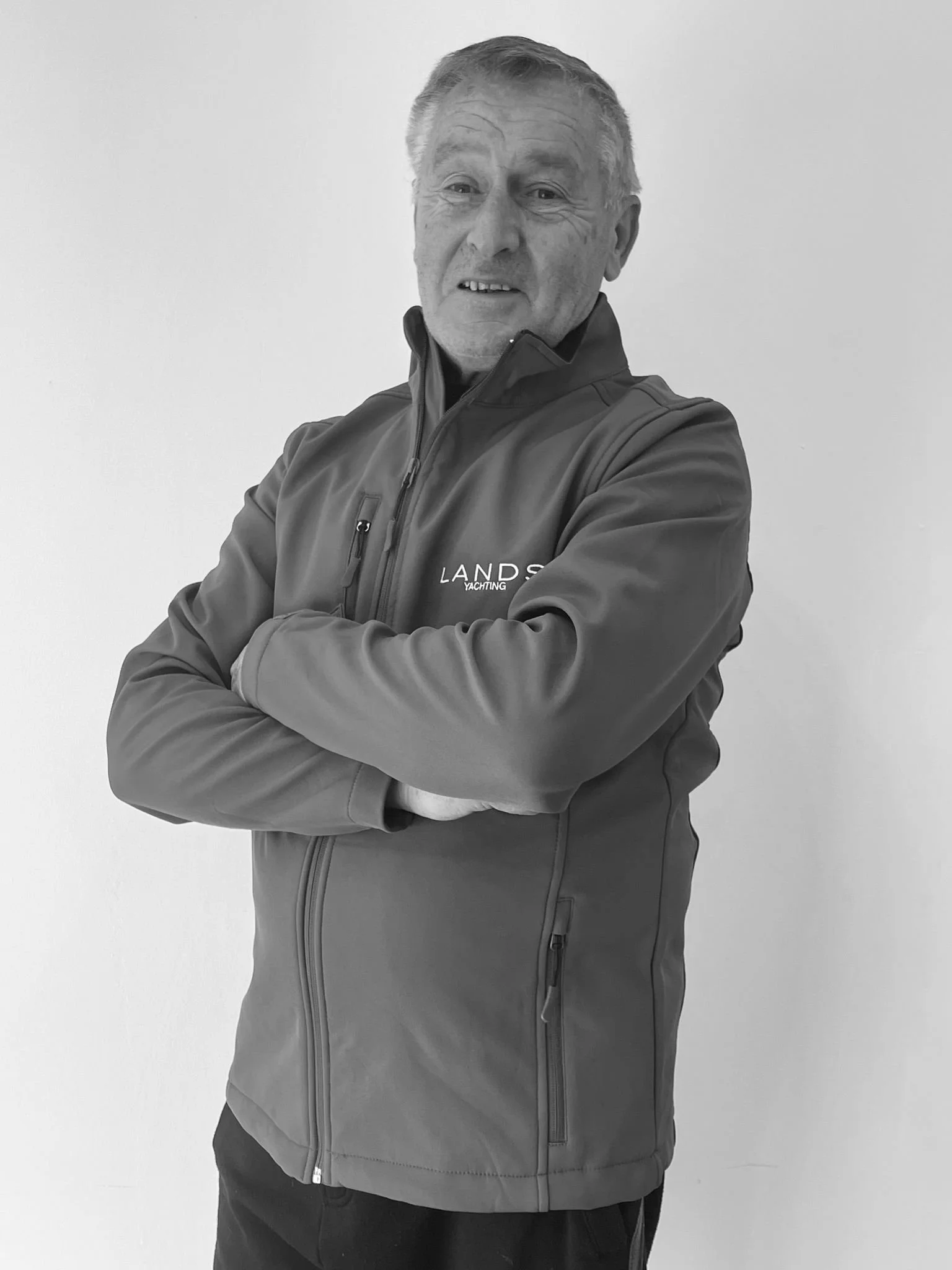 A smiling older man with gray hair wearing a zip-up jacket with his arms crossed, standing in front of a plain white background.