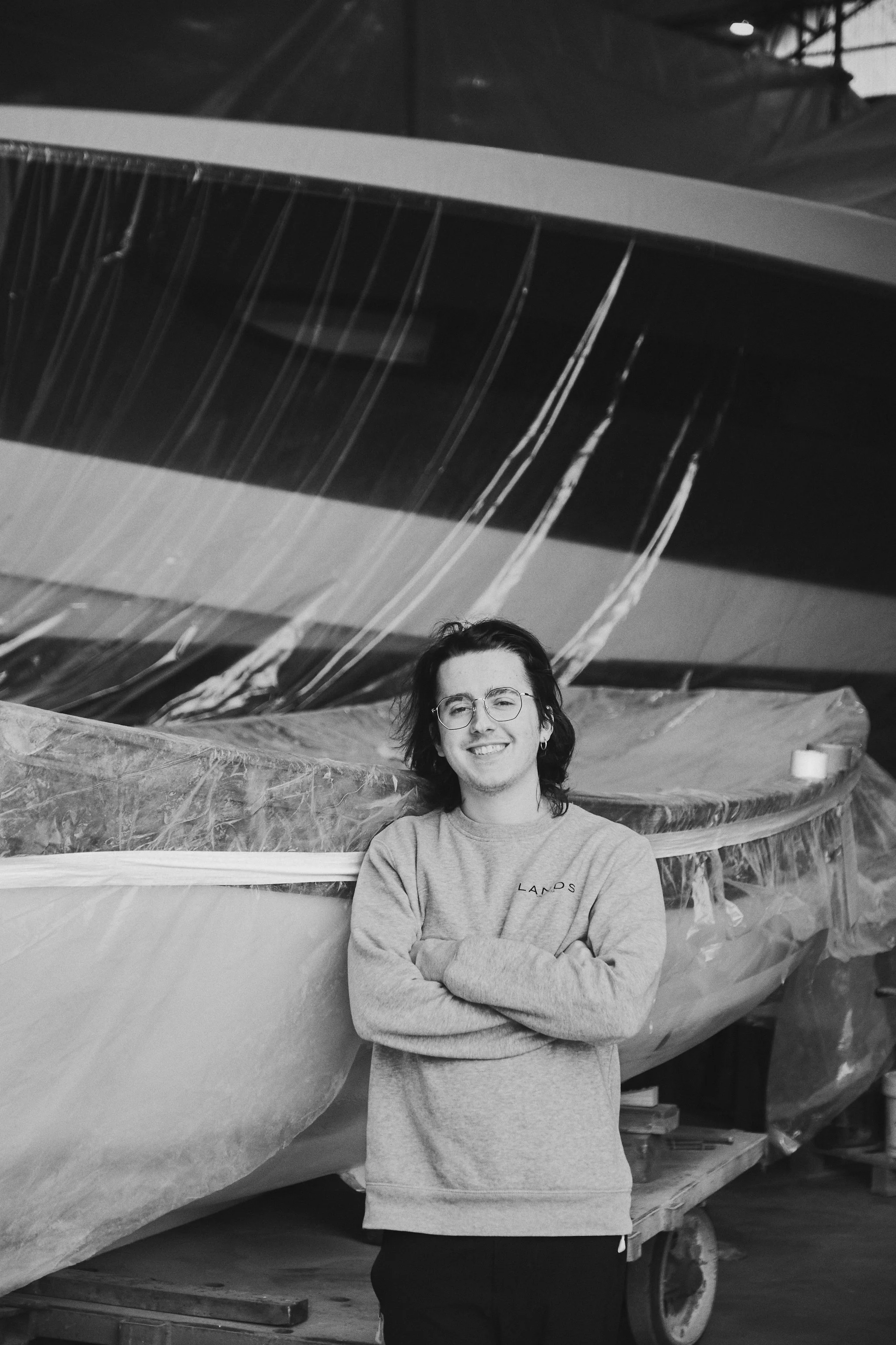 A young man with glasses smiling and crossing his arms, standing in front of a large boat covered in plastic in a boat shop or warehouse.