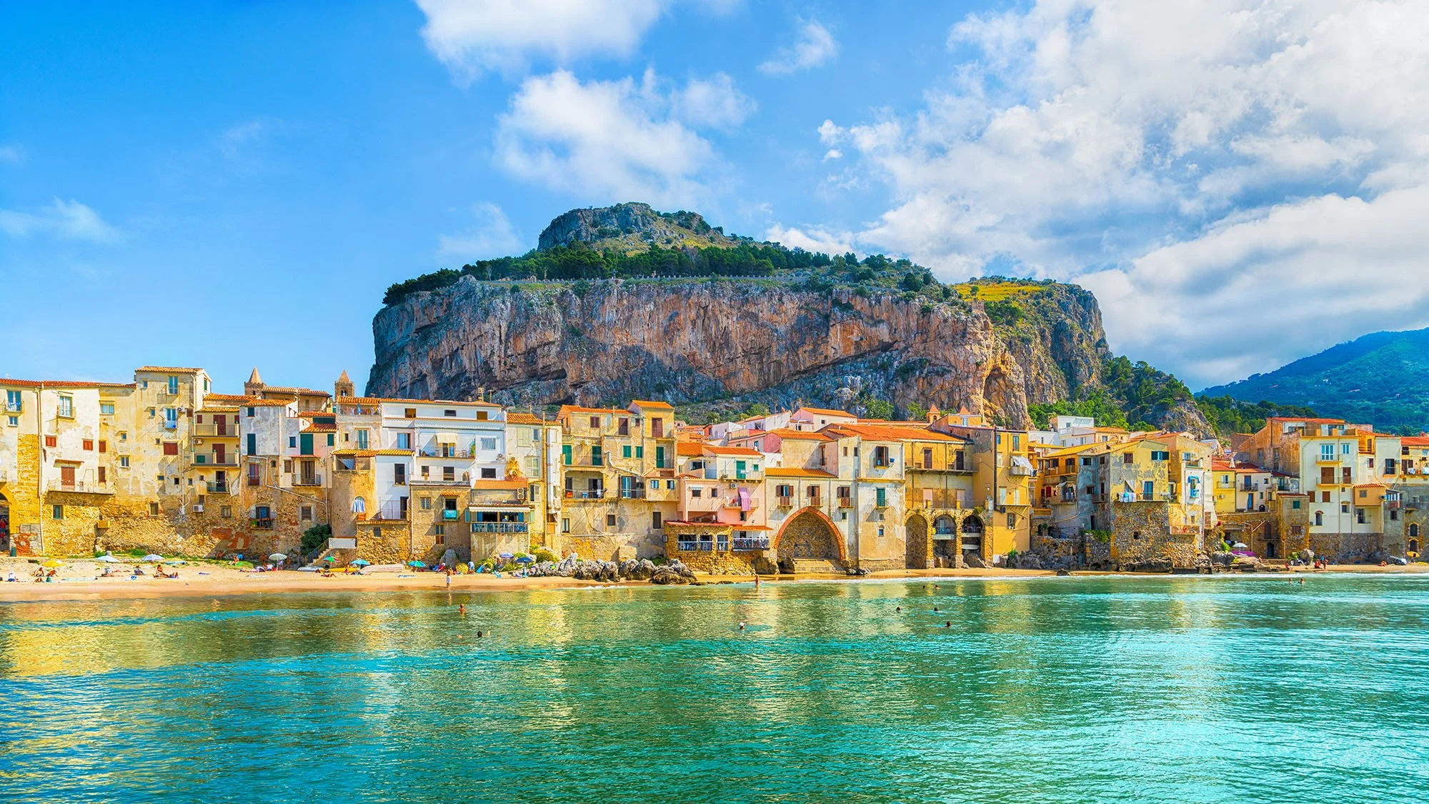Colorful Mediterranean-style buildings along the shoreline with a mountain in the background and a partly cloudy sky.