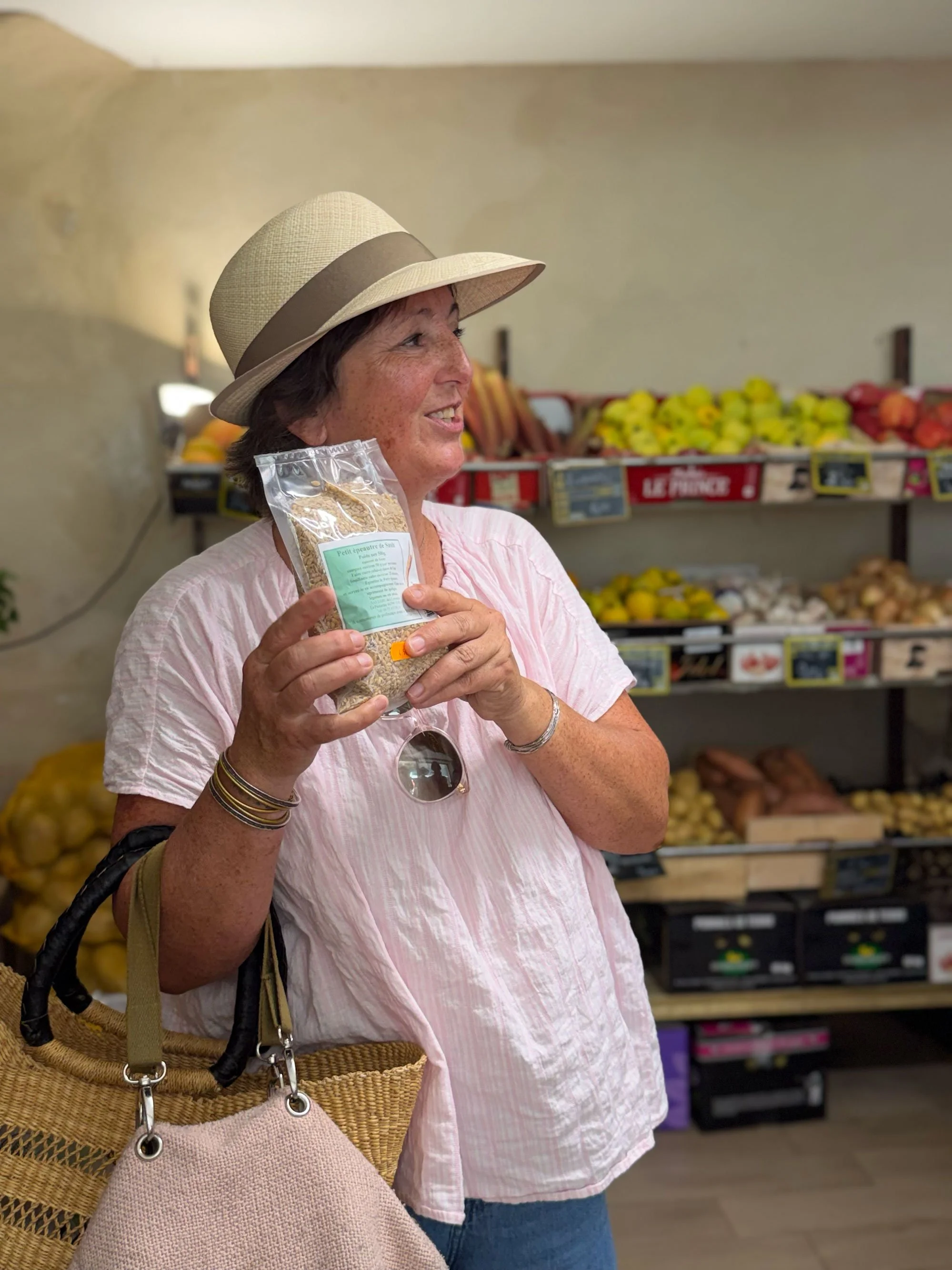 A woman in a wide-brimmed hat, holding a bag of grains in a grocery store with various fruits and vegetables on display in the background.