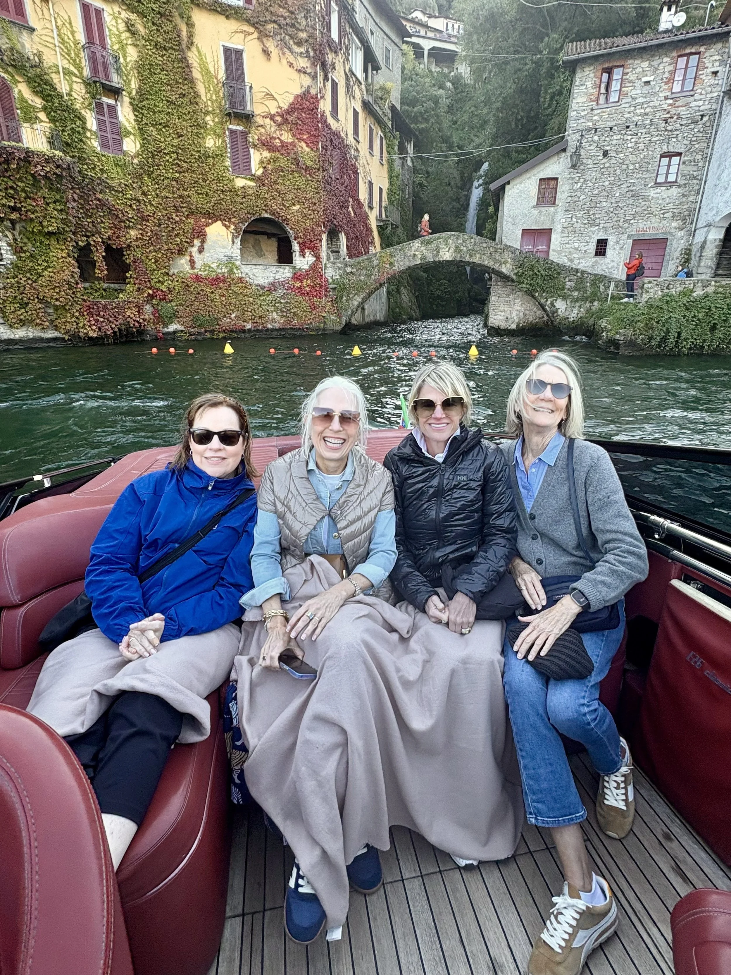 Four women smiling and sitting on a boat with a European village and waterfall in the background.