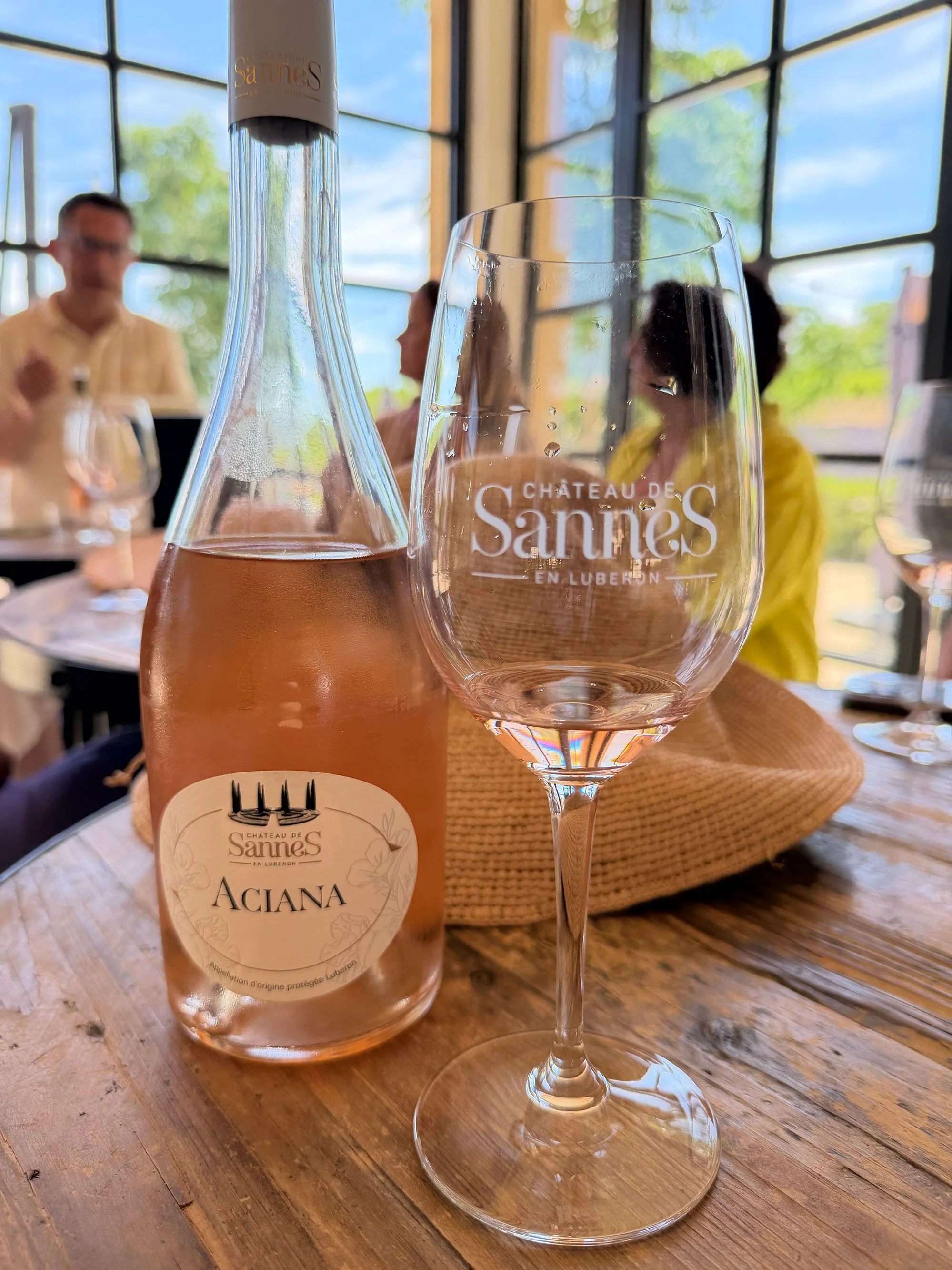 A wine bottle and an empty wine glass on a wooden table, with people sitting in the background inside a glass-walled building on a sunny day.