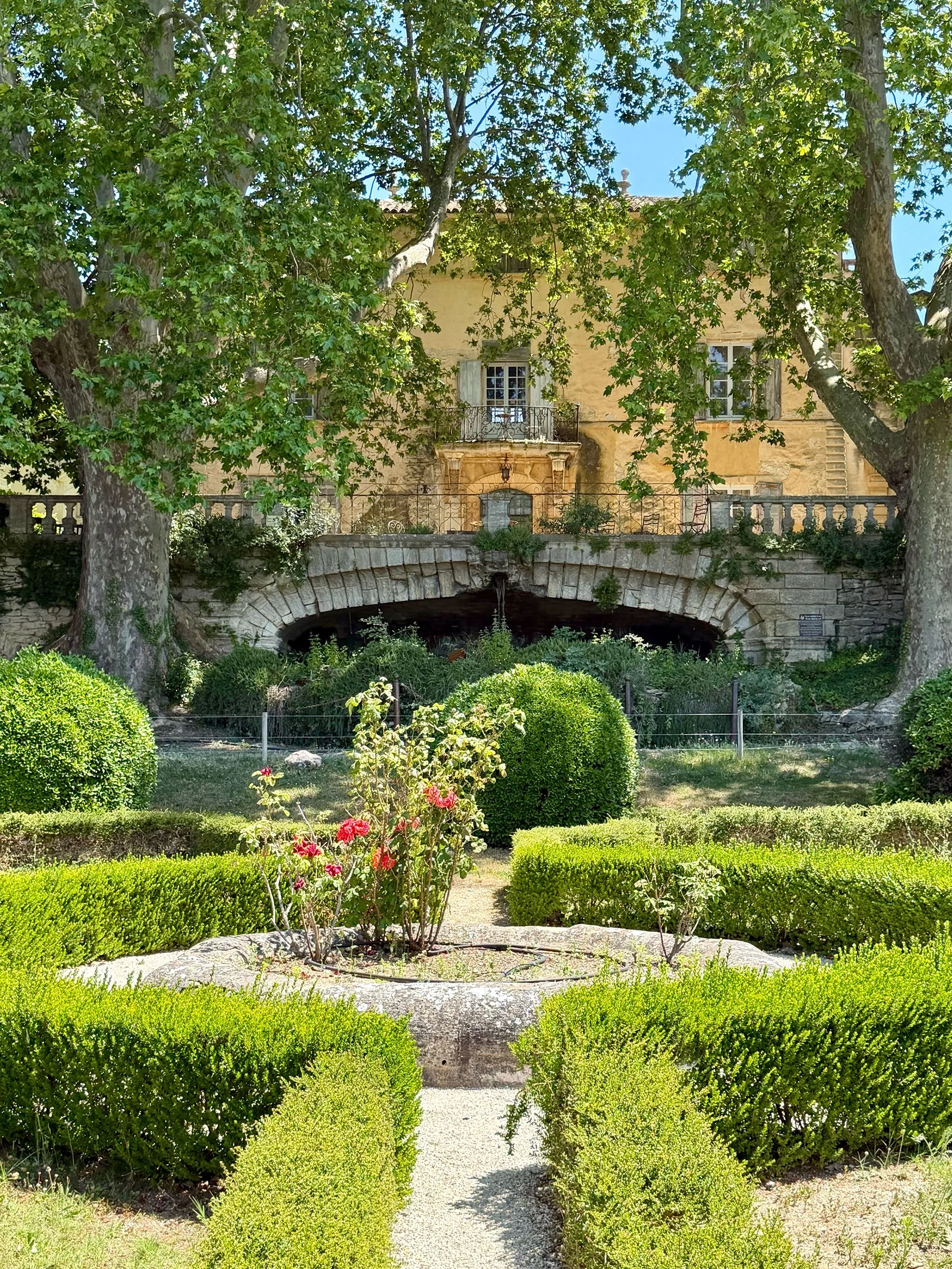 A garden with neatly trimmed green bushes and a stone pathway leading towards an old building with a stone arch and a fountain underneath. Large trees with green leaves partially cover the building.