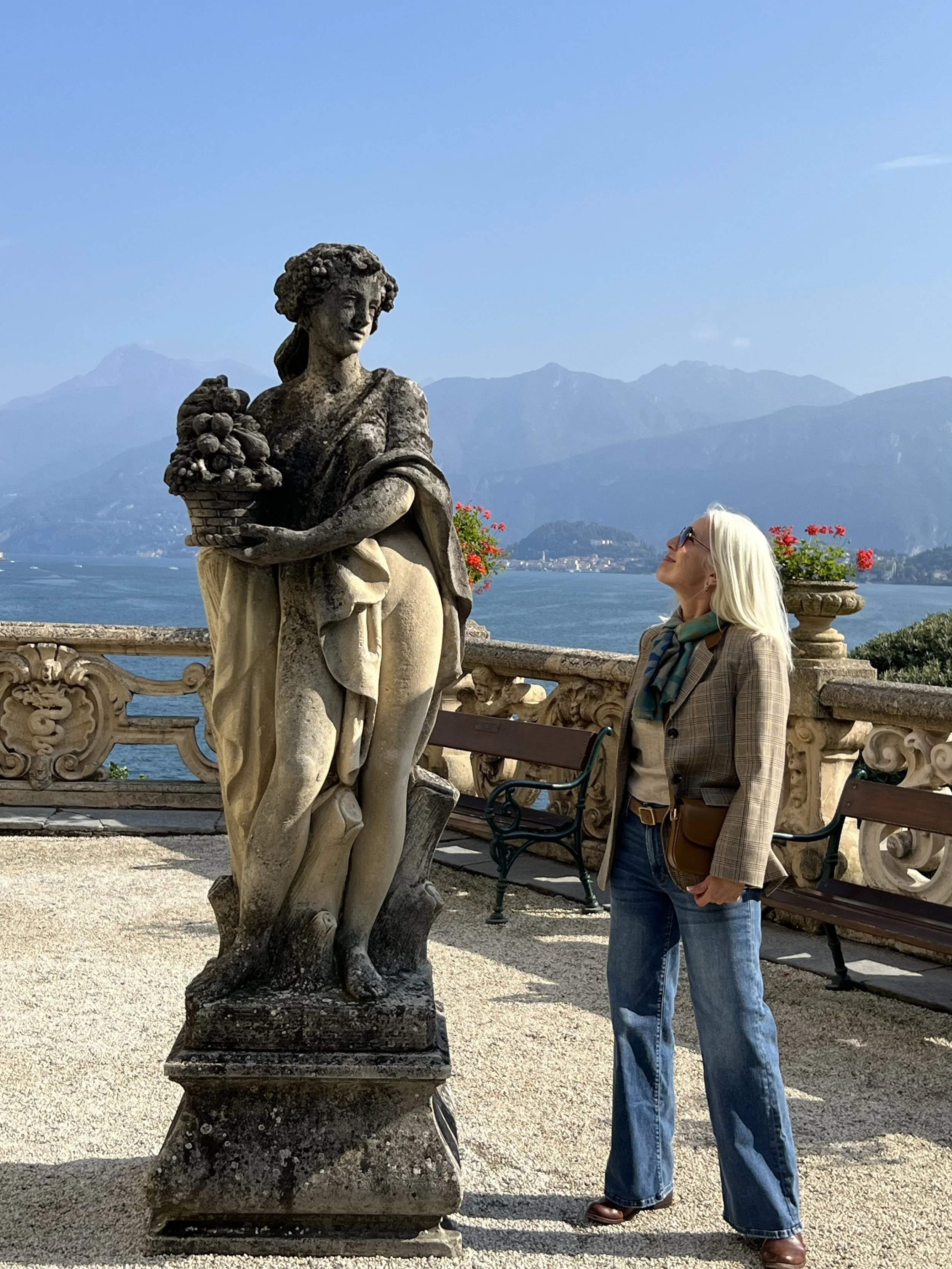 A woman with white hair, sunglasses, plaid blazer, jeans, and a shoulder bag stands next to an old stone statue on a terrace overlooking a lake and mountains. The statue is of a woman holding a basket of fruit.