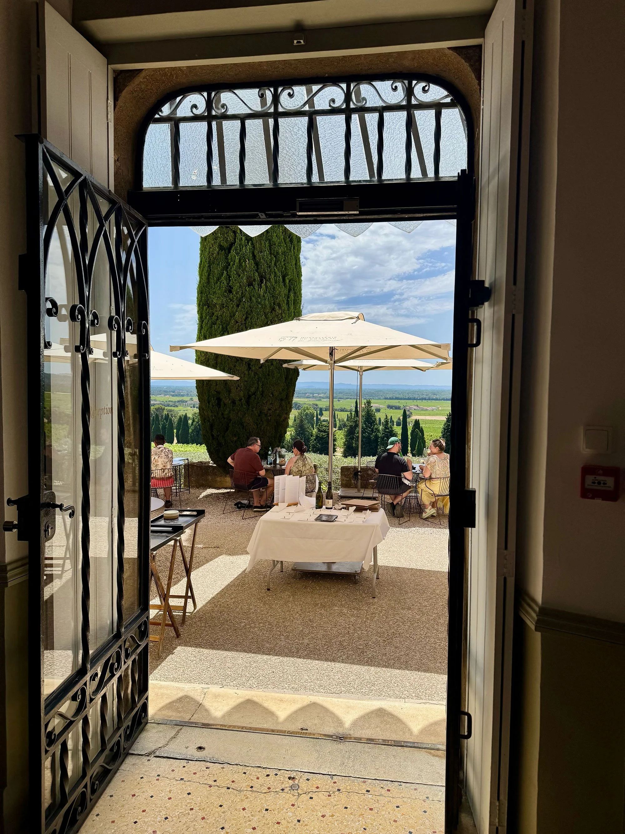 View of an outdoor patio with a large tree, two umbrellas, and people seated, seen through a half-open door with decorative ironwork.