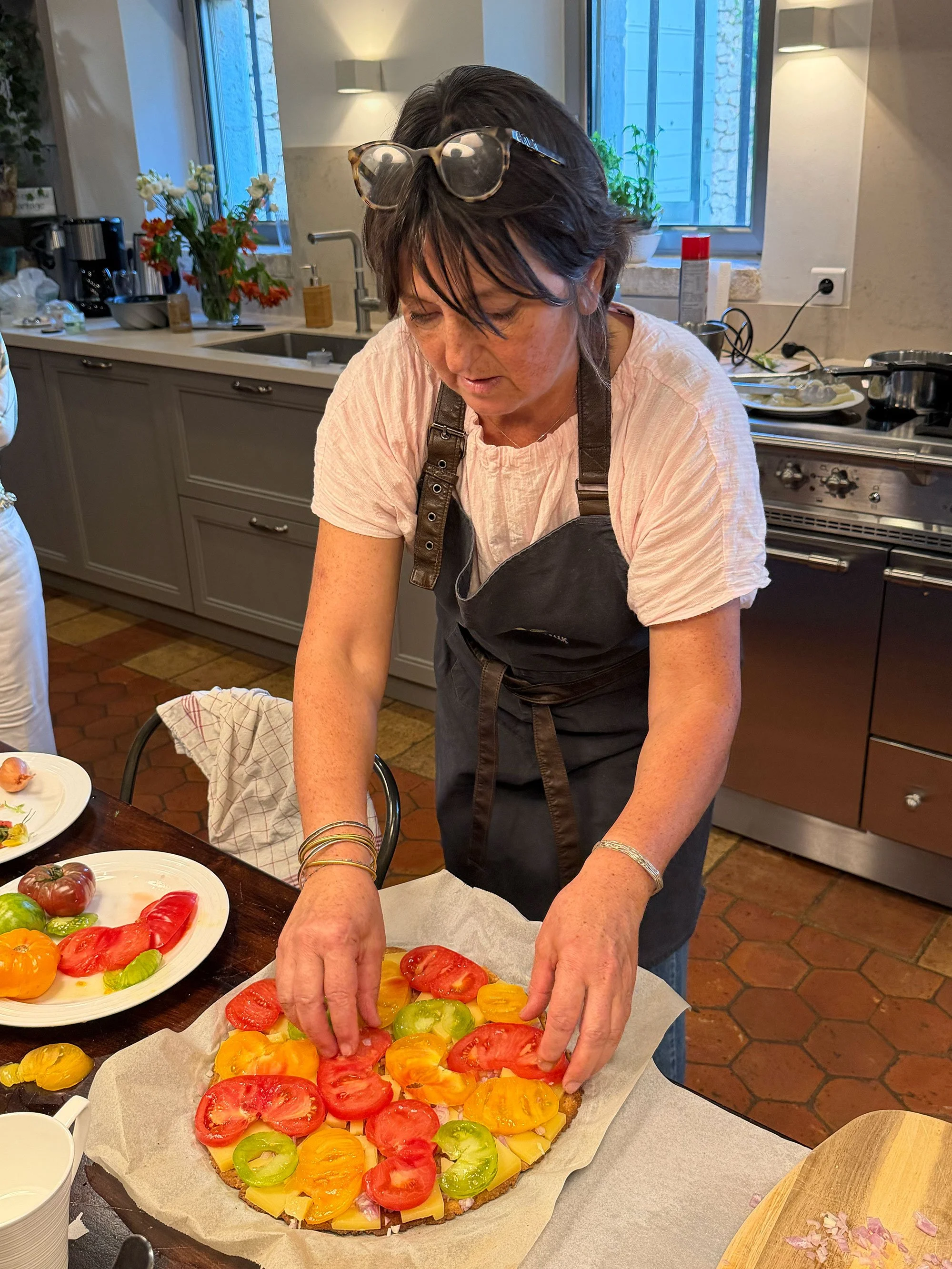 A woman placing sliced tomatoes and yellow bell peppers on a pizza at a kitchen counter.