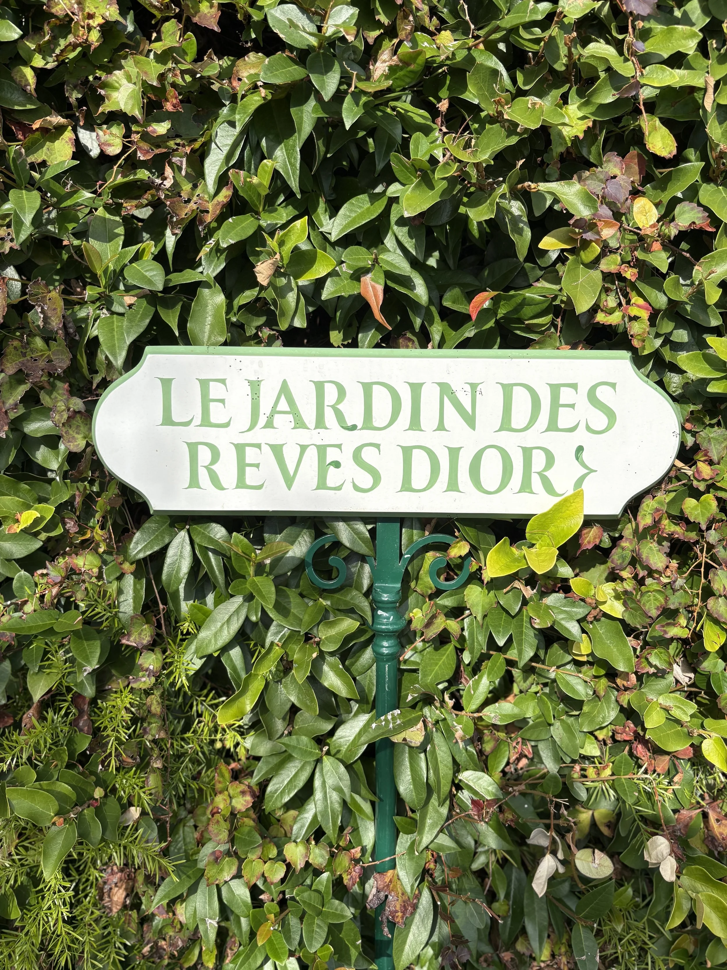 A white garden sign with green text that reads "Le Jardin des Rêves Dior," mounted on a green metal stake, positioned in front of lush green hedges and foliage.