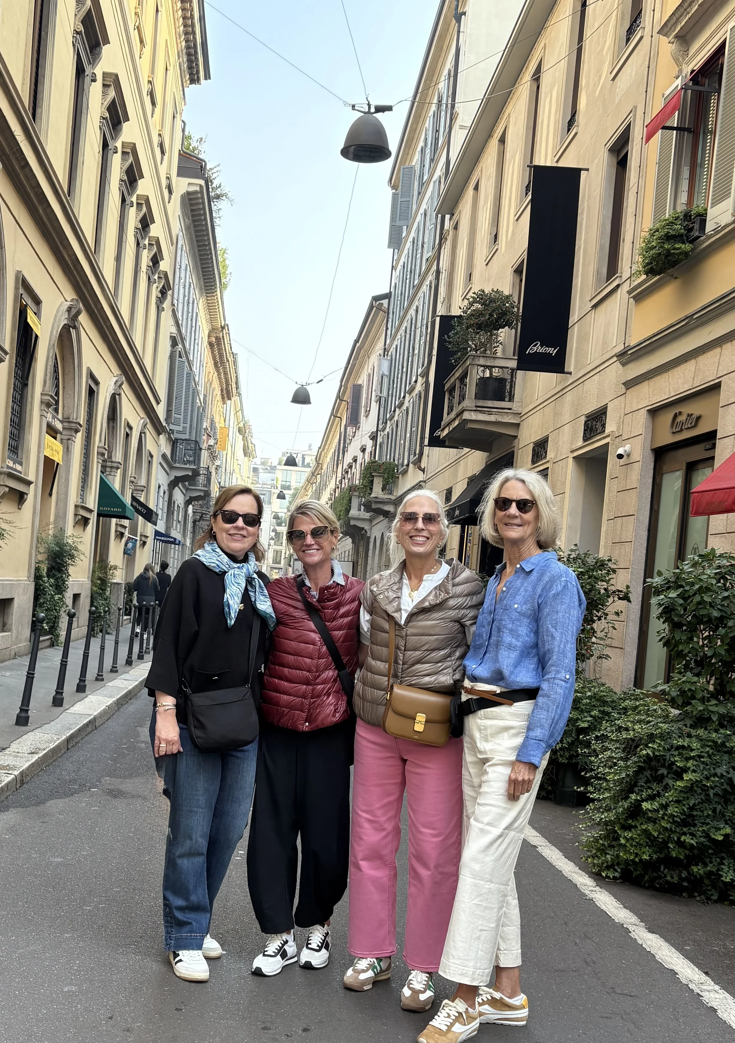 Four women standing close together in a European city street, smiling at the camera, wearing casual clothing and sunglasses.