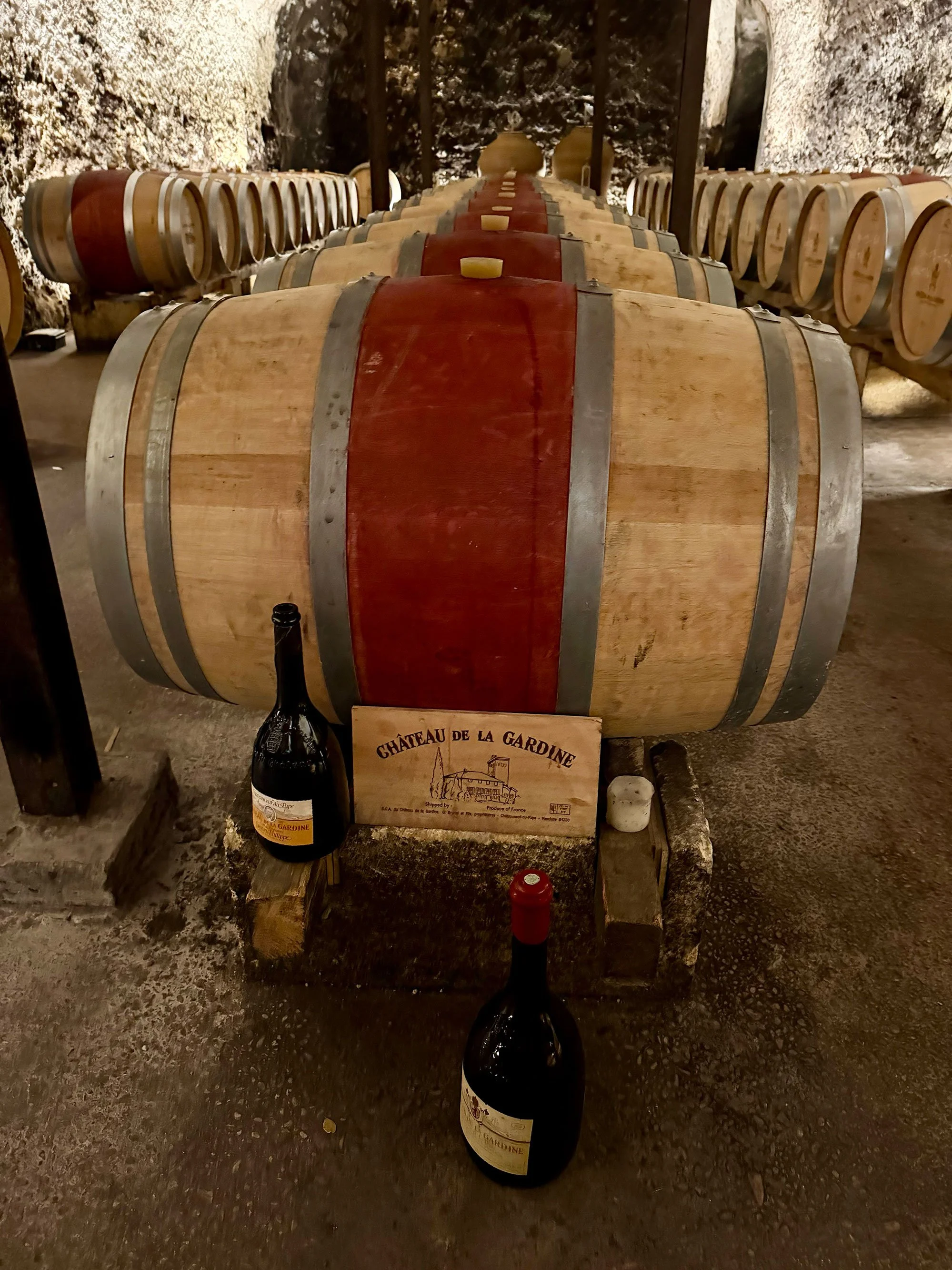 A collection of wooden wine barrels in a wine cellar, with a sign reading 'Château de la Gardine', two wine bottles, a candle, and a small stone on the ground in front.