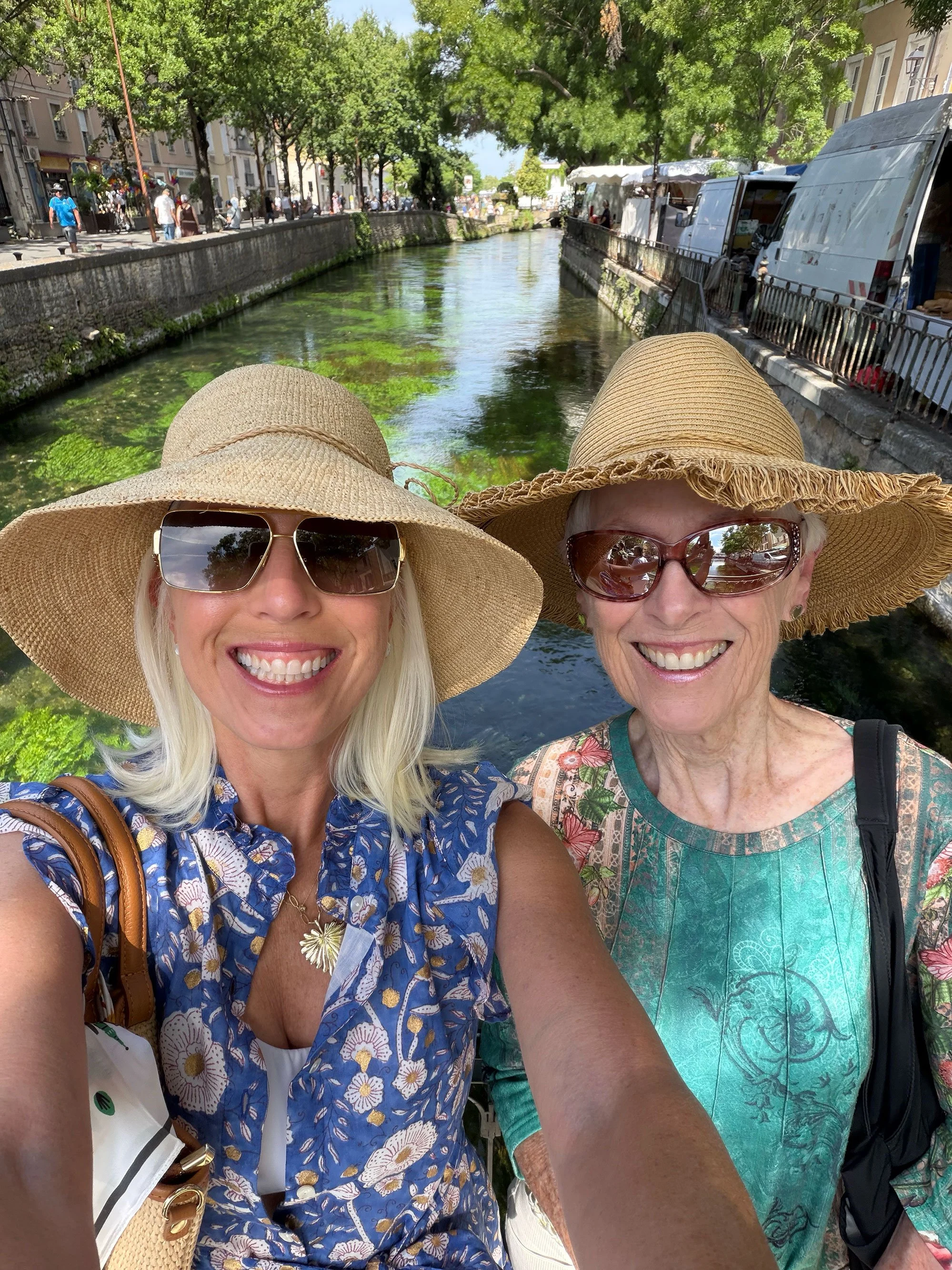 Two women wearing wide-brimmed straw hats and sunglasses taking a selfie beside a canal with trees and buildings in the background.