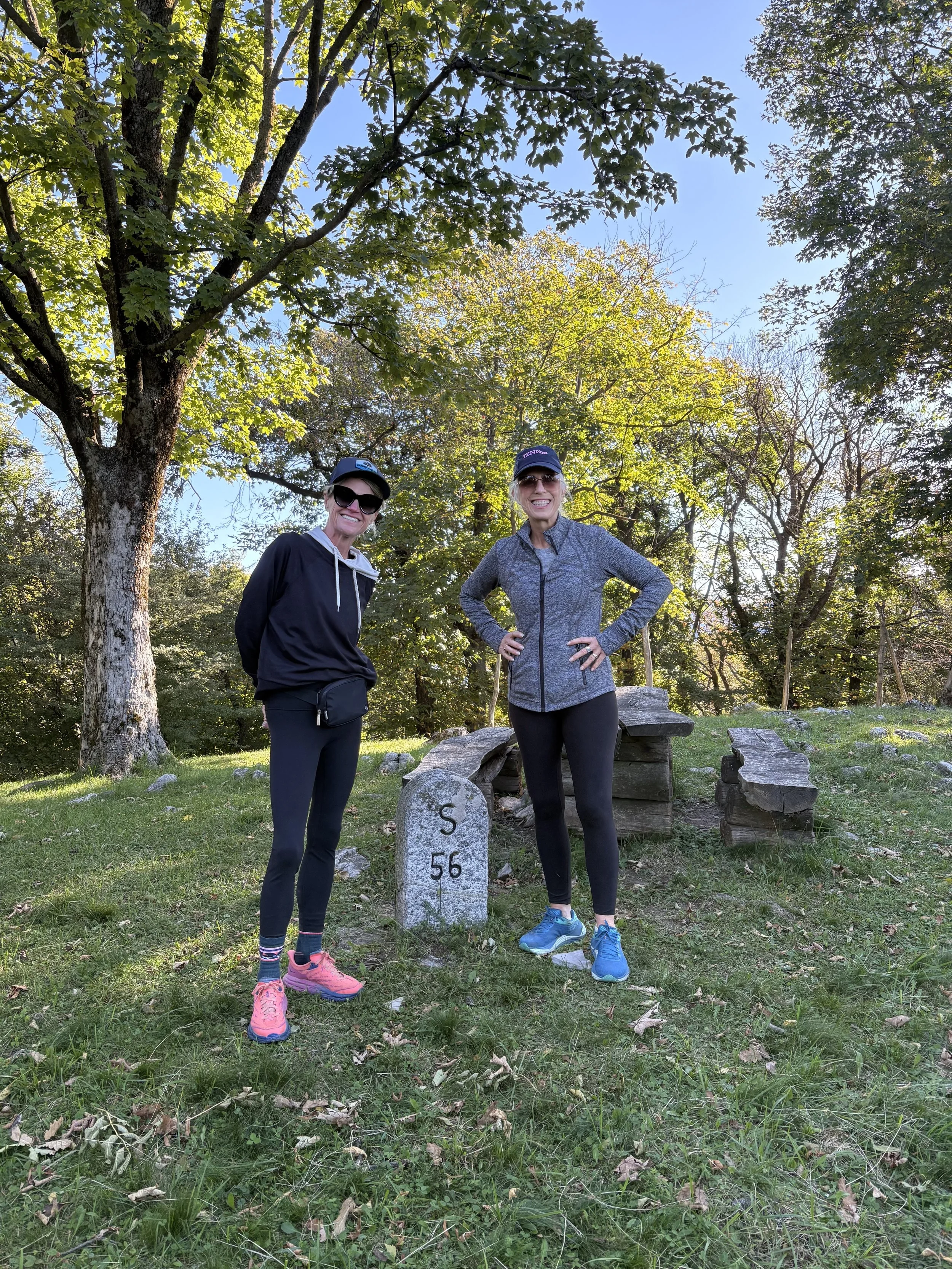 Two women standing outdoors in a forested area, smiling, wearing athletic clothing and sunglasses. They are posing next to a stone marker with the numbers 'S 56' engraved on it, with trees and a bright blue sky in the background.