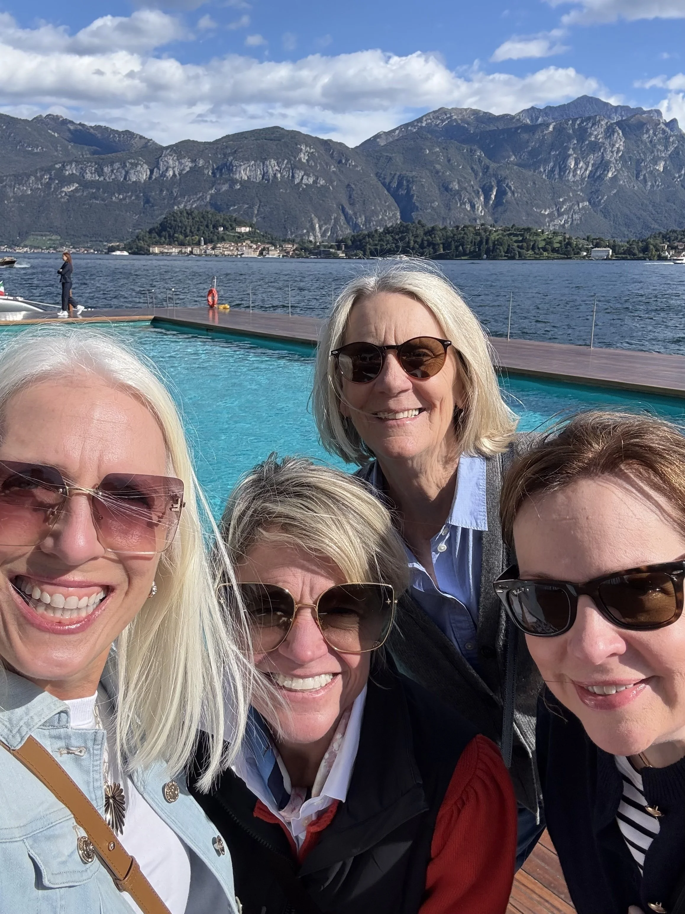 Group of five women smiling outdoors near a swimming pool with a mountain lake and mountains in the background.