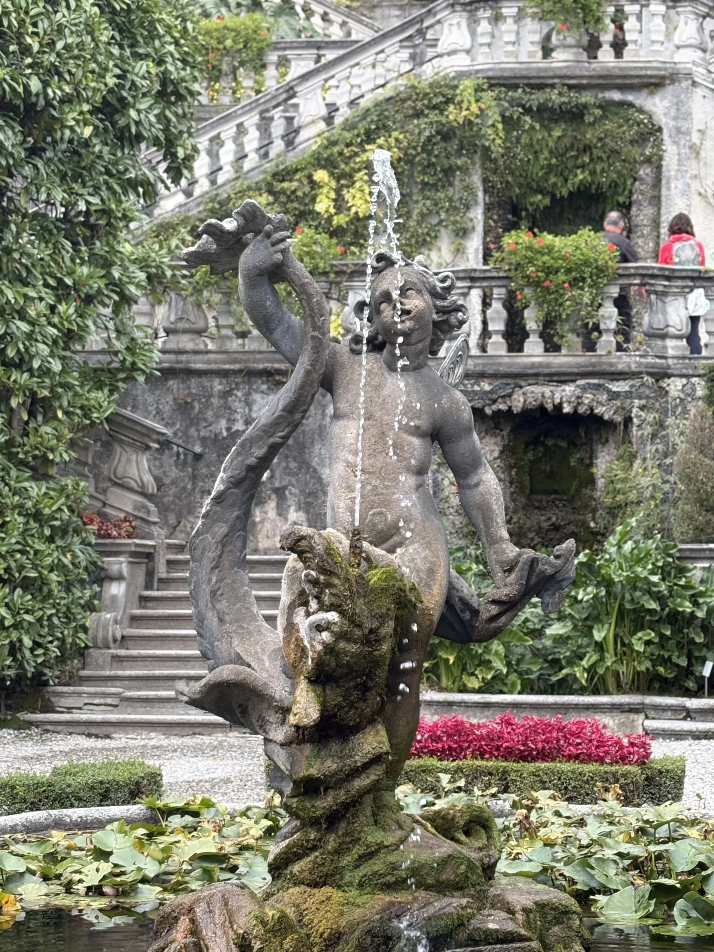 Statue of a mythological figure with water spouting from its head, surrounded by greenery and flowers, in a garden setting.