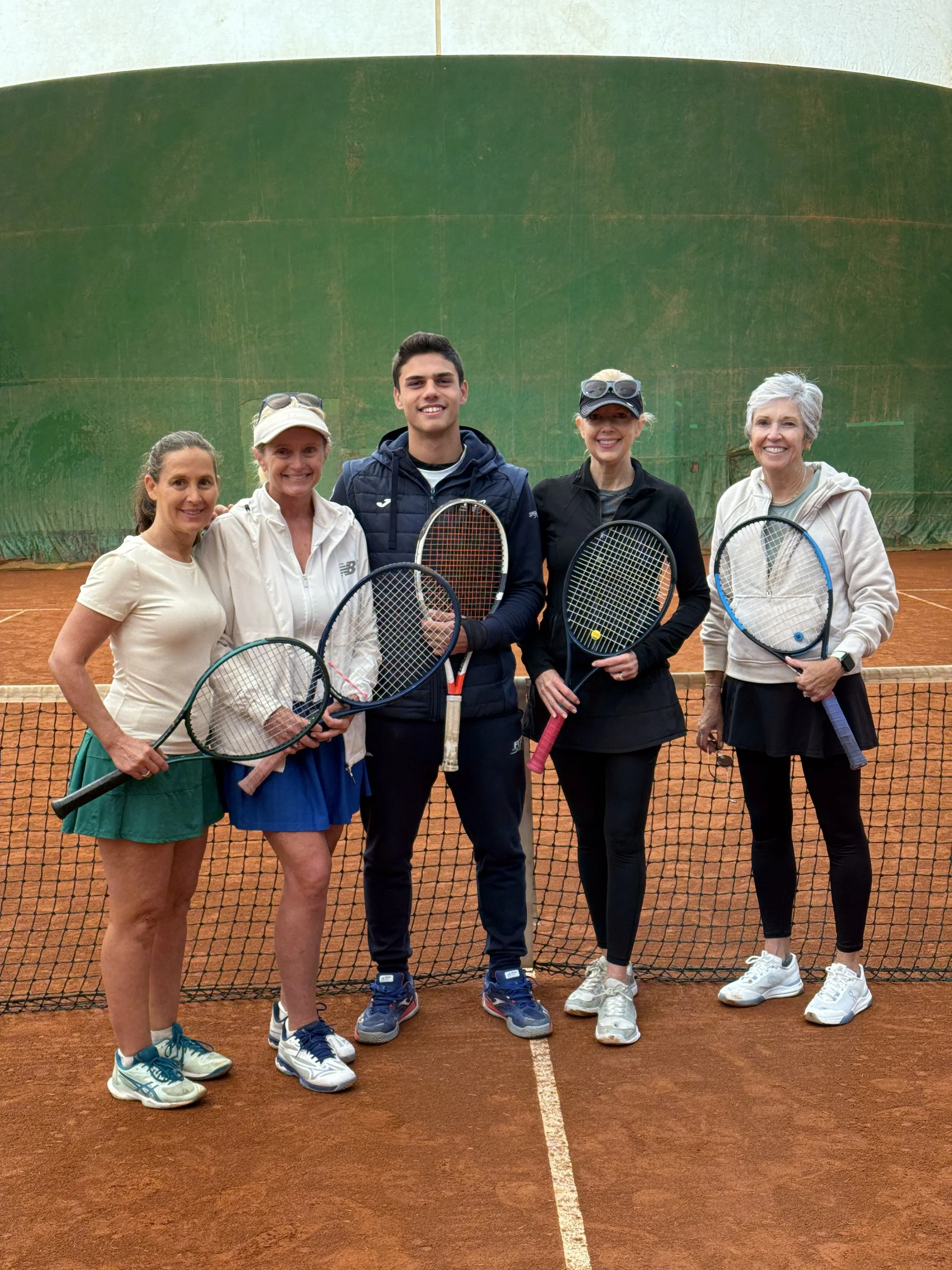Group of five people standing on a tennis court holding tennis rackets, smiling, with a large green wall in the background.