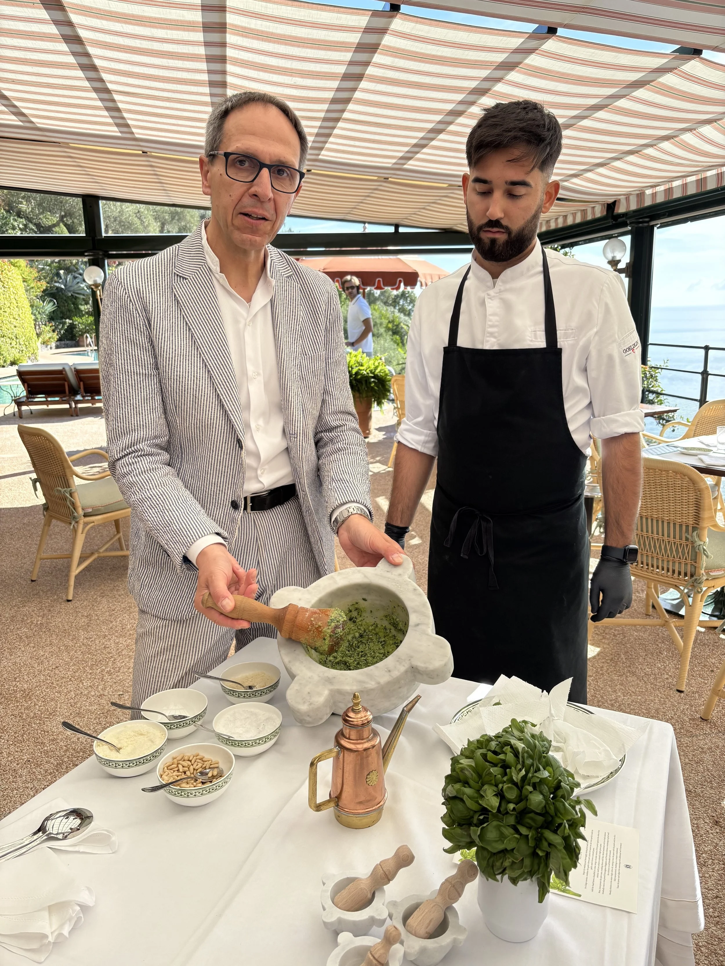 A man in a striped suit demonstrating how to prepare pesto with a mortar and pestle, accompanied by a chef in black apron at a table with bowls of ingredients, in an outdoor restaurant setting.