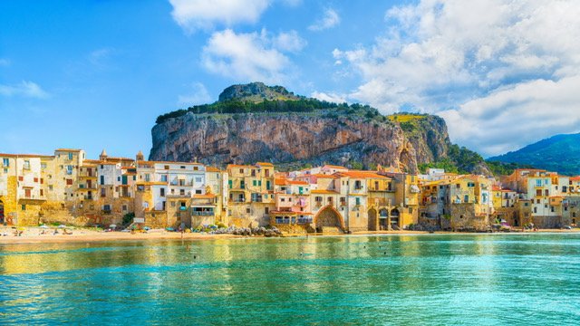 Colorful coastal village with buildings along the water, a large rocky hill behind, and a partly cloudy sky in Verdura, Taormina & Palermo.