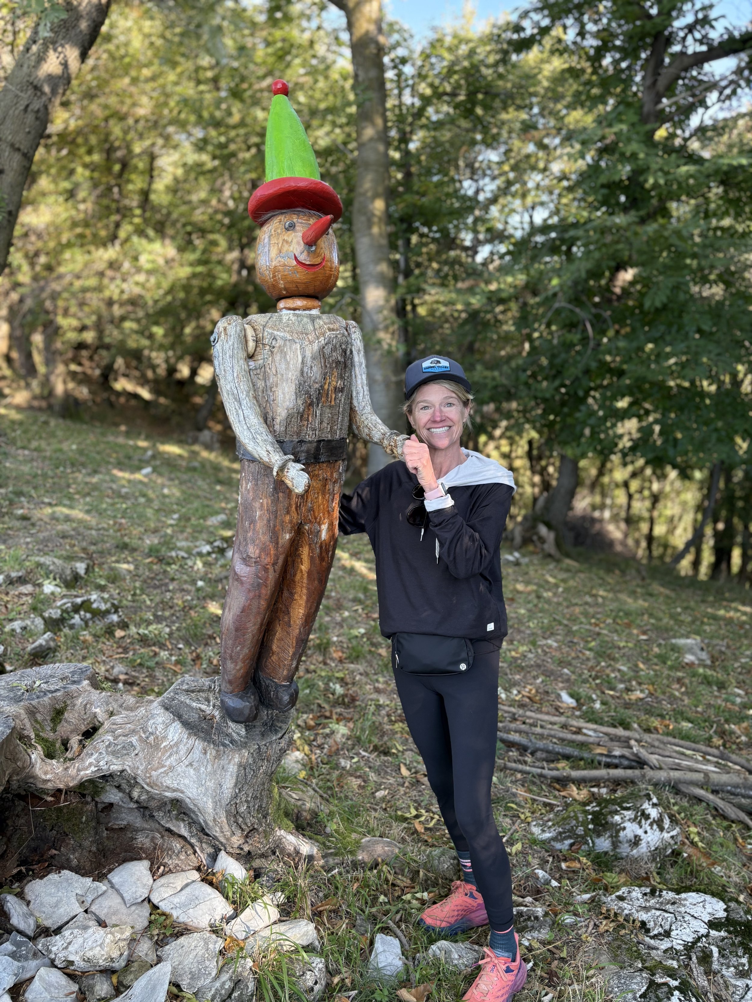 A woman standing in a wooded area next to a tall wooden sculpture of a clown, holding its hand and smiling at the camera.