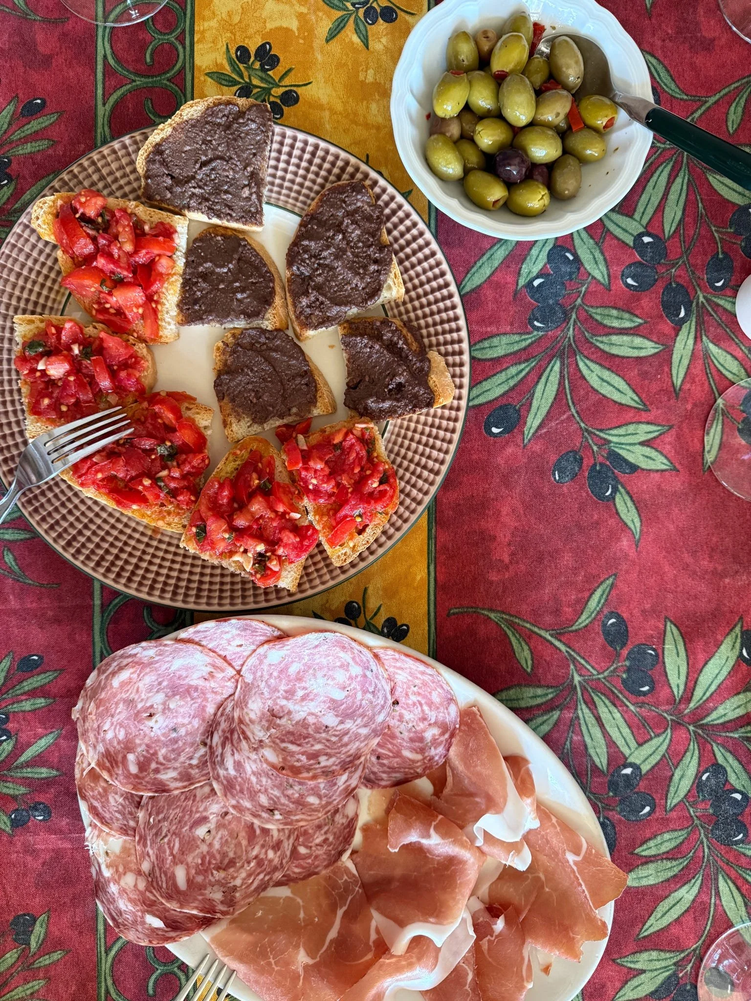 Plate of sliced cured meats, including salami and prosciutto, on a table with a red, green, and yellow olive-themed tablecloth. Also, a bowl of green and black olives and a plate of bruschetta with tomato and basil topping.