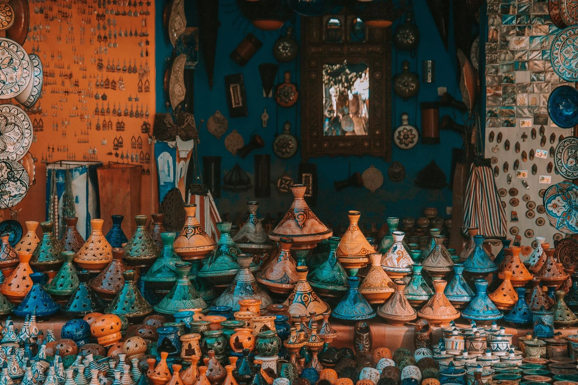 A marketplace stall displaying a variety of colorful traditional pottery, including centuries-old clay vessels and decorative items, set against a vibrant blue and orange backdrop with hanging artwork and accessories.