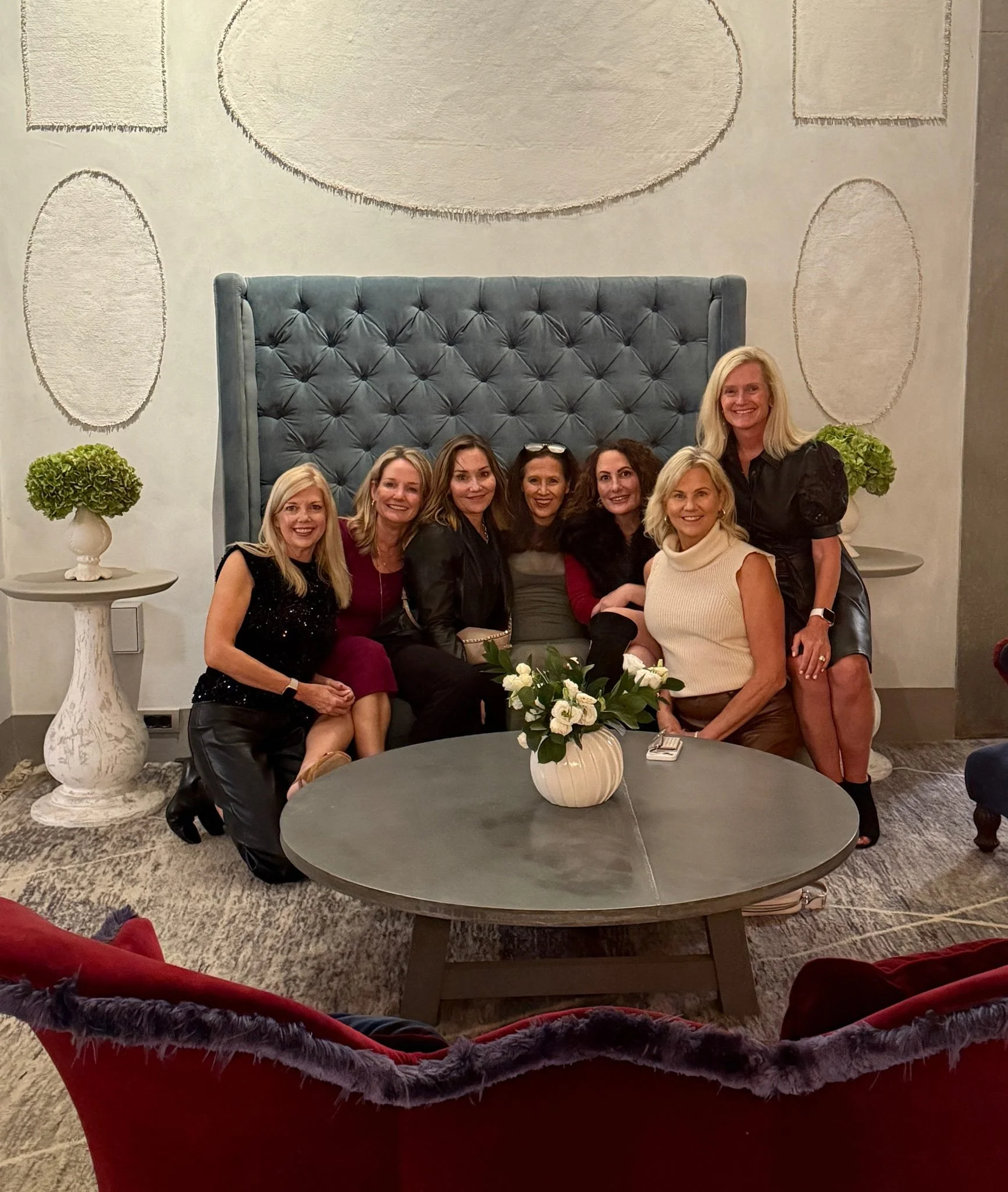 Group of nine women sitting and kneeling on and around a sectional sofa in a well-decorated room, smiling at the camera.