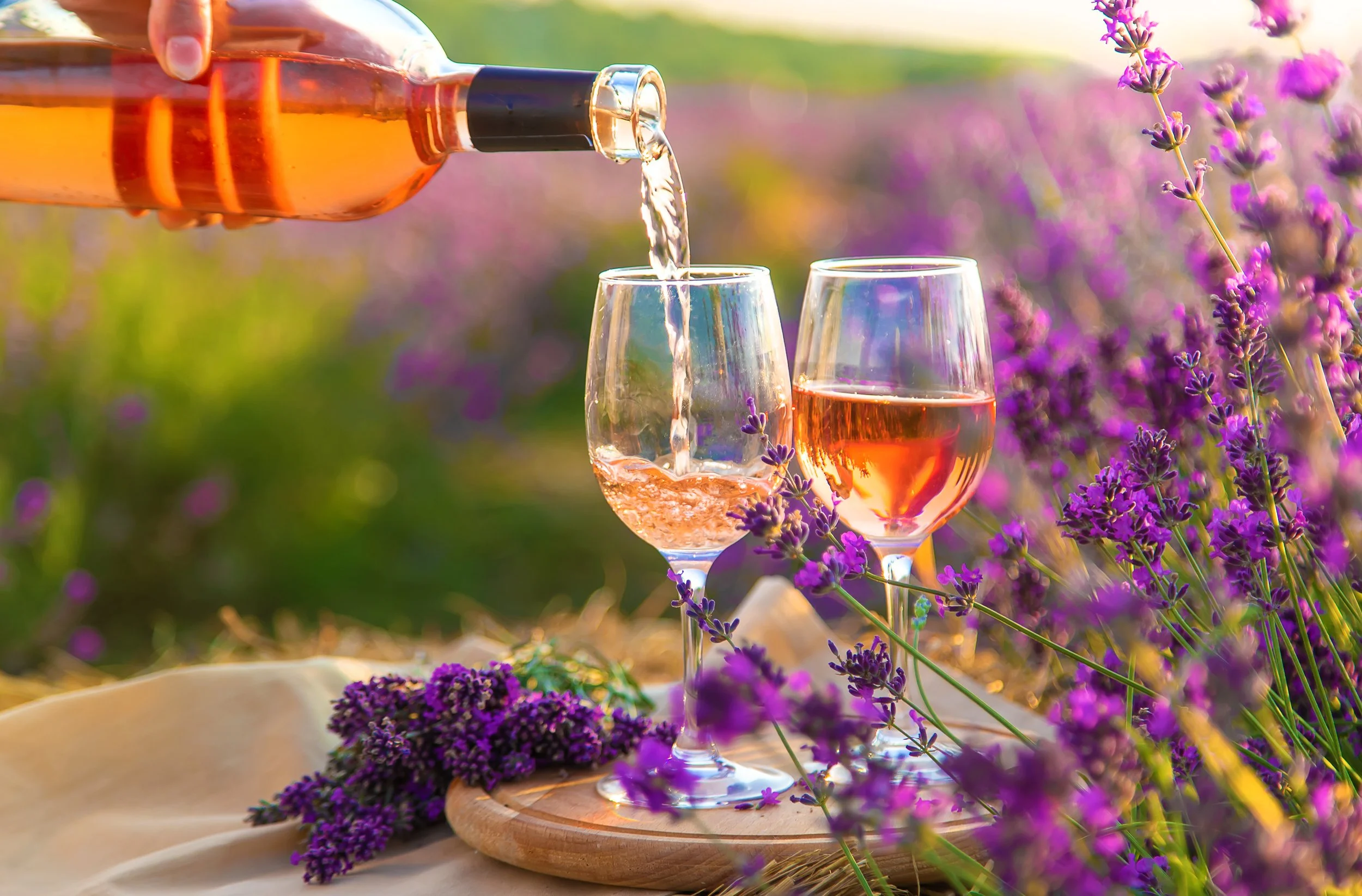 A person pours rosé wine into a wine glass outdoors with purple lavender flowers and a blurred garden background.