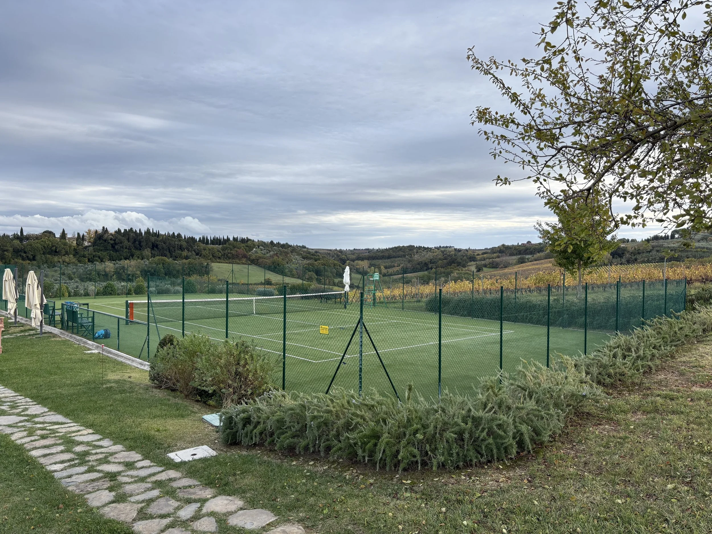 Empty outdoor tennis court surrounded by a green fence, with grey clouds over a landscape of rolling hills and vineyards.