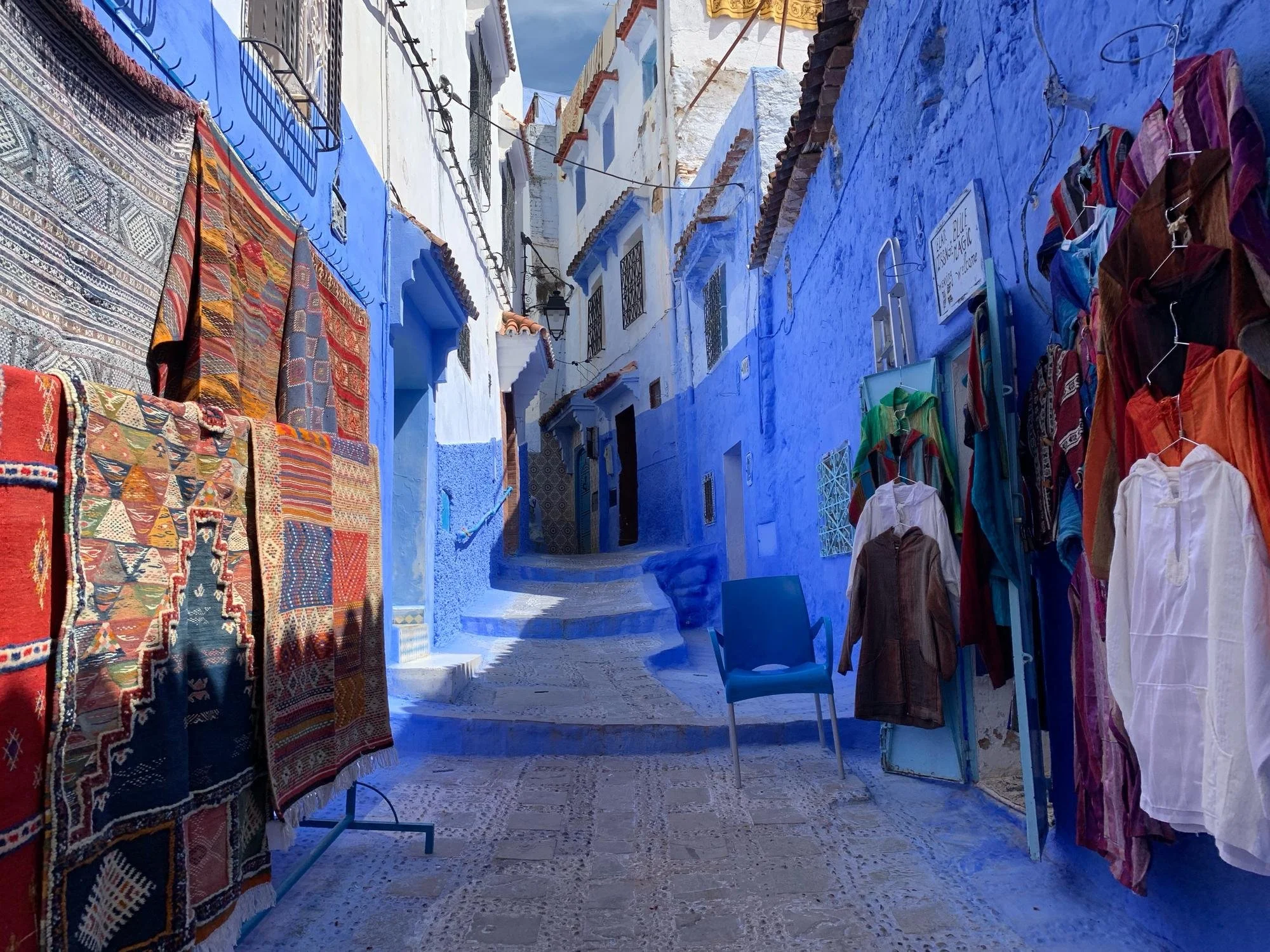 Outdoor market stall with colorful rugs and clothing hanging on racks, white and blue painted buildings in the background.