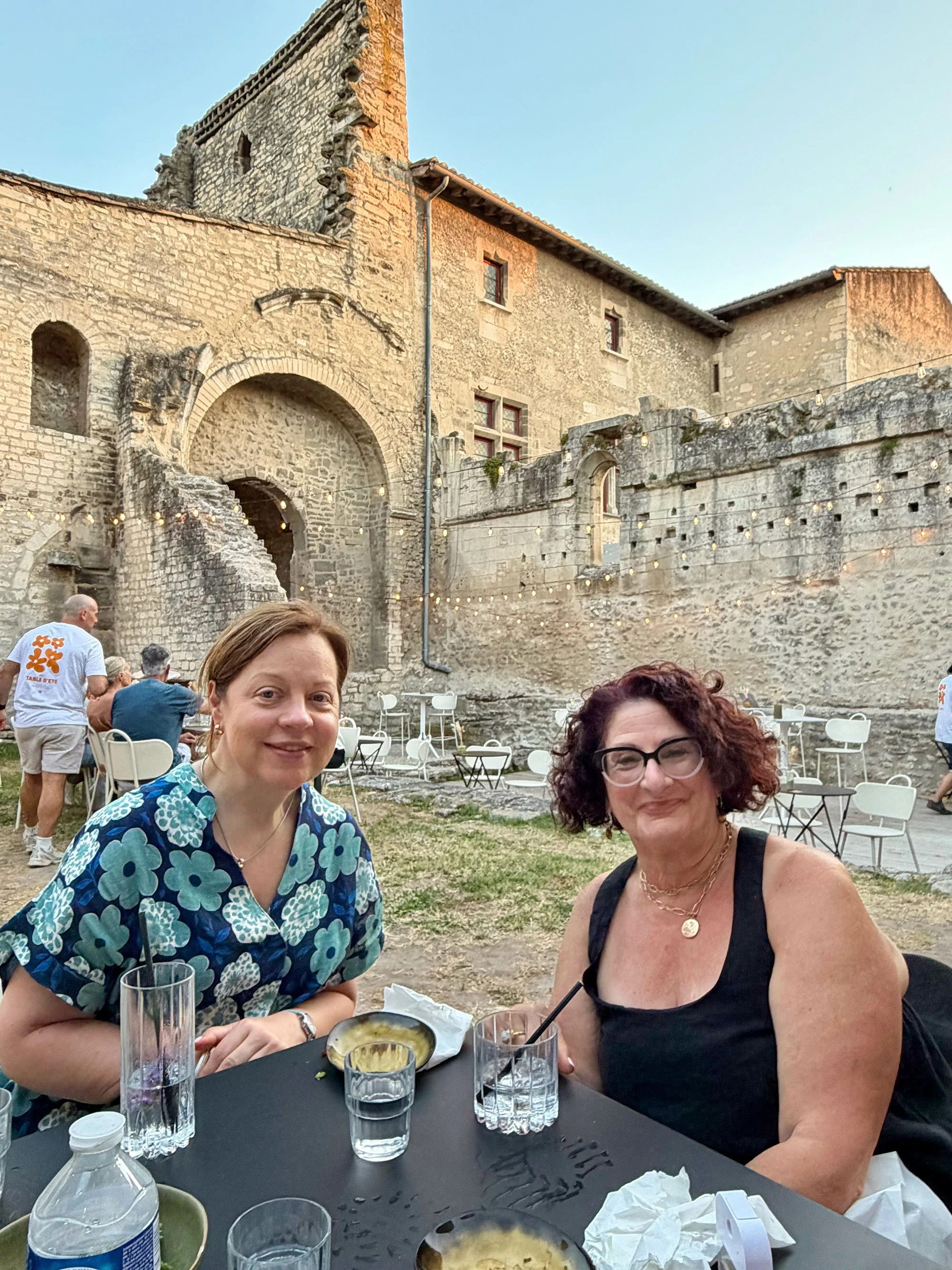 Two women sitting at an outdoor table in front of an old stone building with string lights, enjoying drinks and food.