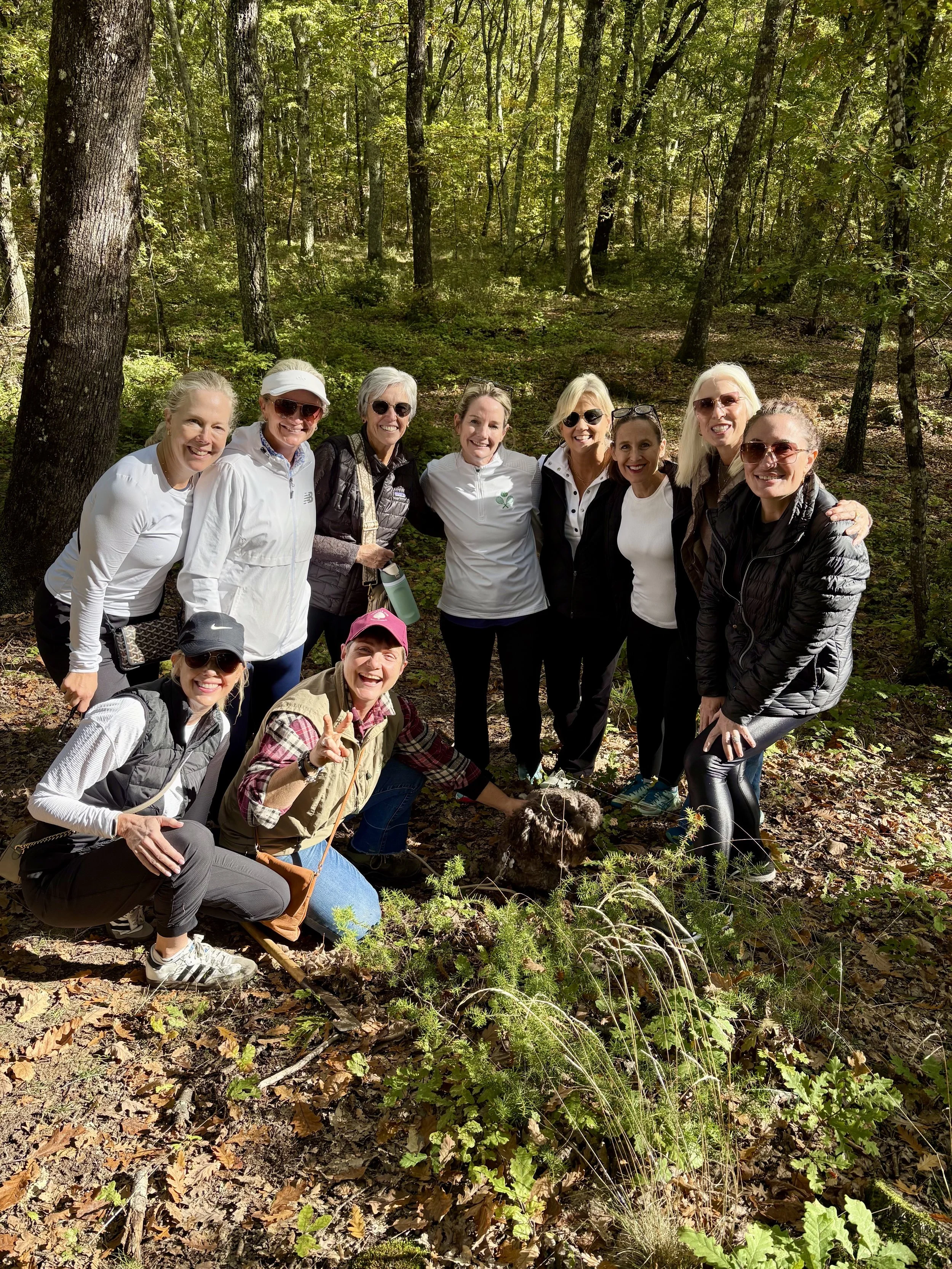 Group of women in a forest during the daytime posing for a photo, some smiling and one woman making a peace sign, with a small furry animal or object on the ground in front of them.