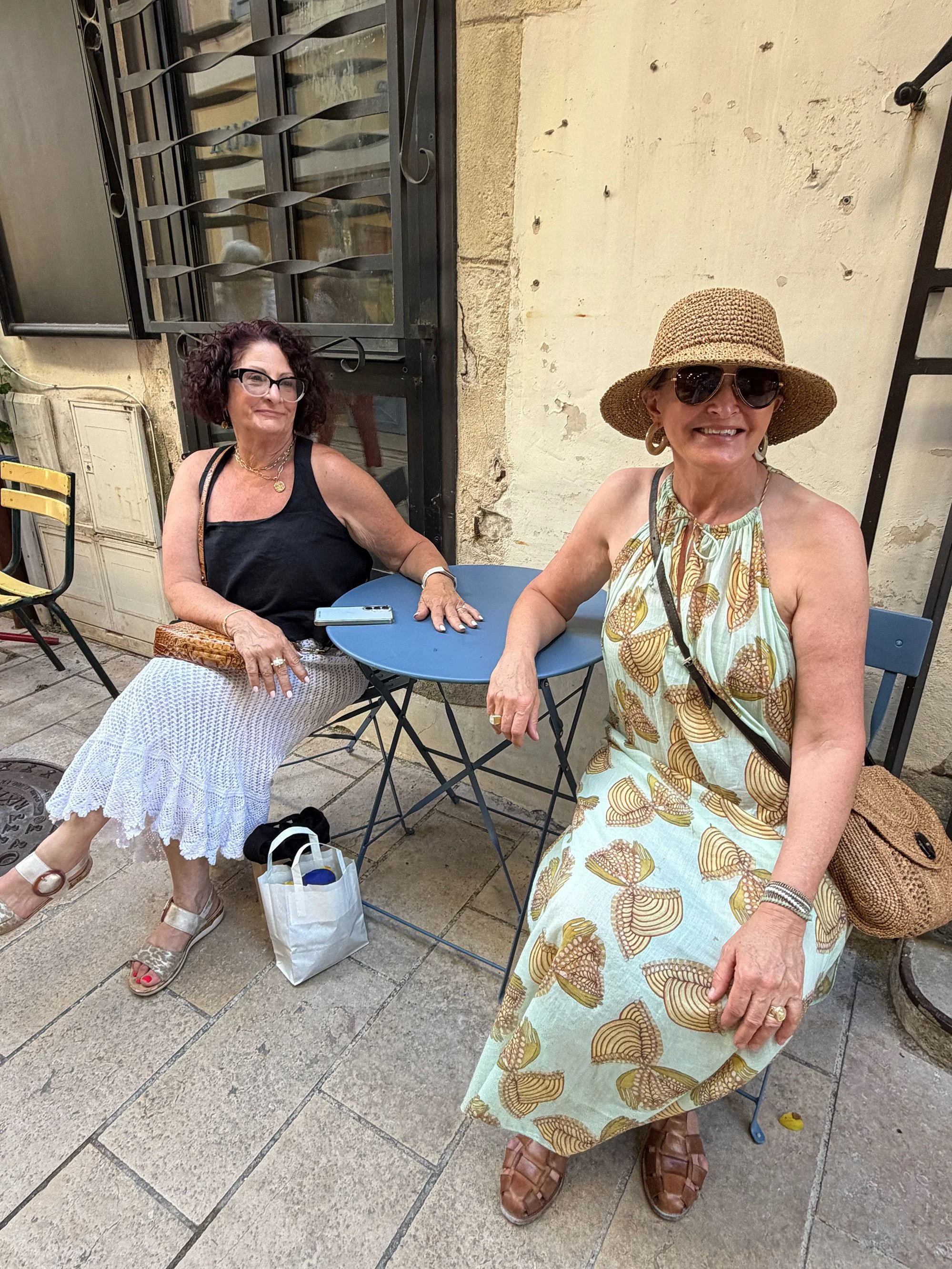 Two women sitting at a blue metal outdoor table. One woman is wearing a black sleeveless top, glasses, white netted skirt, and sandals, with her bag on the ground and a phone on the table. The other woman is wearing a wide-brimmed straw hat, sunglass
