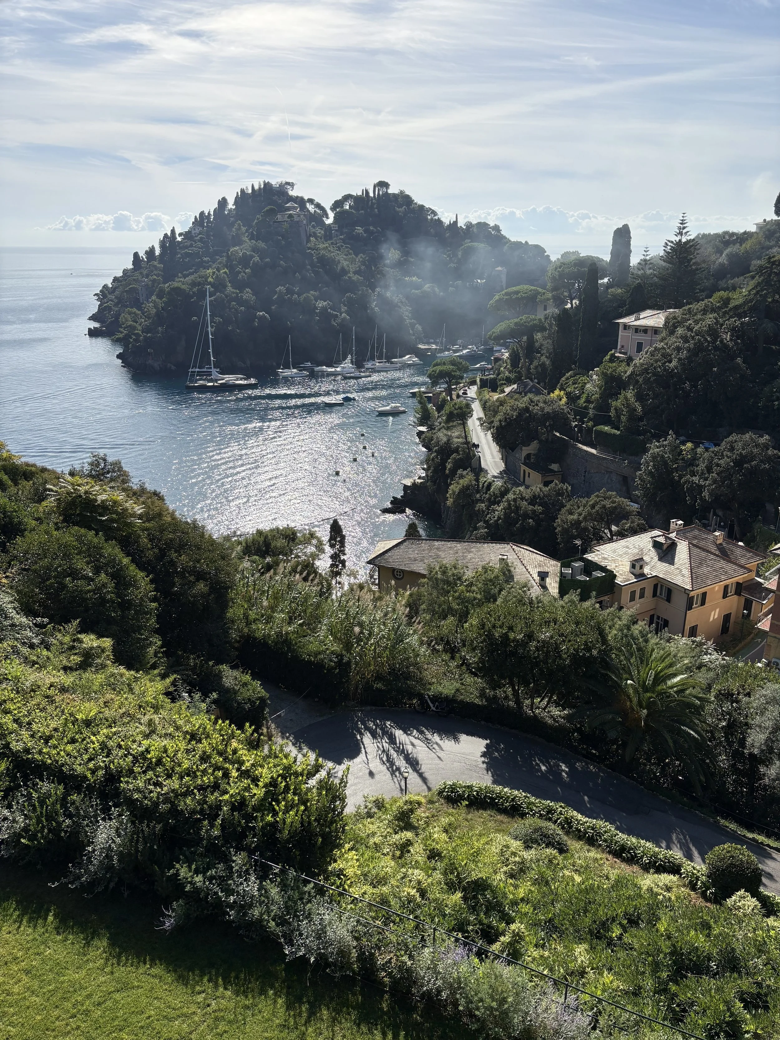 Scenic view of a coastal hillside with houses, lush greenery, and a bay with sailboats, under a partly cloudy sky.